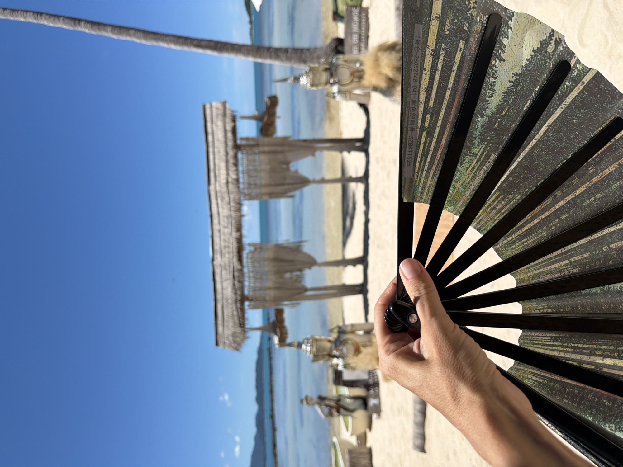 The image shows a person's hand holding a black folding fan, with the fan's patterned side facing the viewer. The fan is partially obscuring a tropical beach scene. In the background, there is a thatched-roof structure, a palm tree, and the ocean under a clear blue sky. The water appears calm, and the sand is light-colored. The scene suggests a warm, sunny day, likely in the late morning or early afternoon given the bright sunlight and shadows. The overall mood is relaxed and peaceful, fitting for a beach setting. There is some text visible on the fan, but it is mostly obscured and difficult to read. The location context provided is Baan Thong Krut, Thailand, which aligns with the tropical beach scenery.