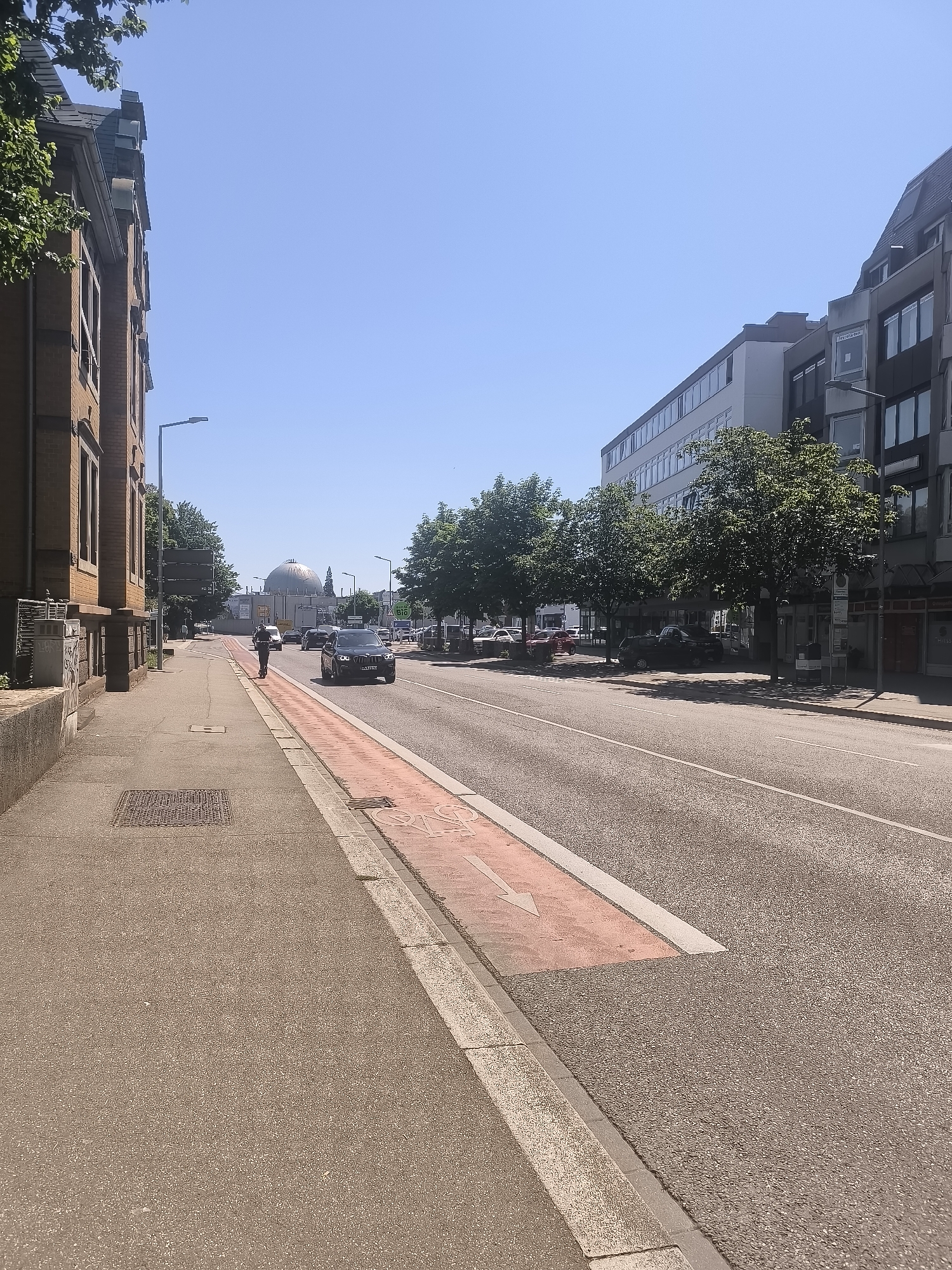 The image shows a street scene in a city, with buildings on the left and right sides and a road in the center. The sky is clear and blue. On the left, a brick building with several windows is visible, next to a sidewalk and a bike lane. A person on a scooter is riding on the sidewalk. The road has multiple lanes of traffic, with several cars driving. In the background, a large, domed building is visible. Trees line the street on both sides. The scene appears to be on a sunny day.
