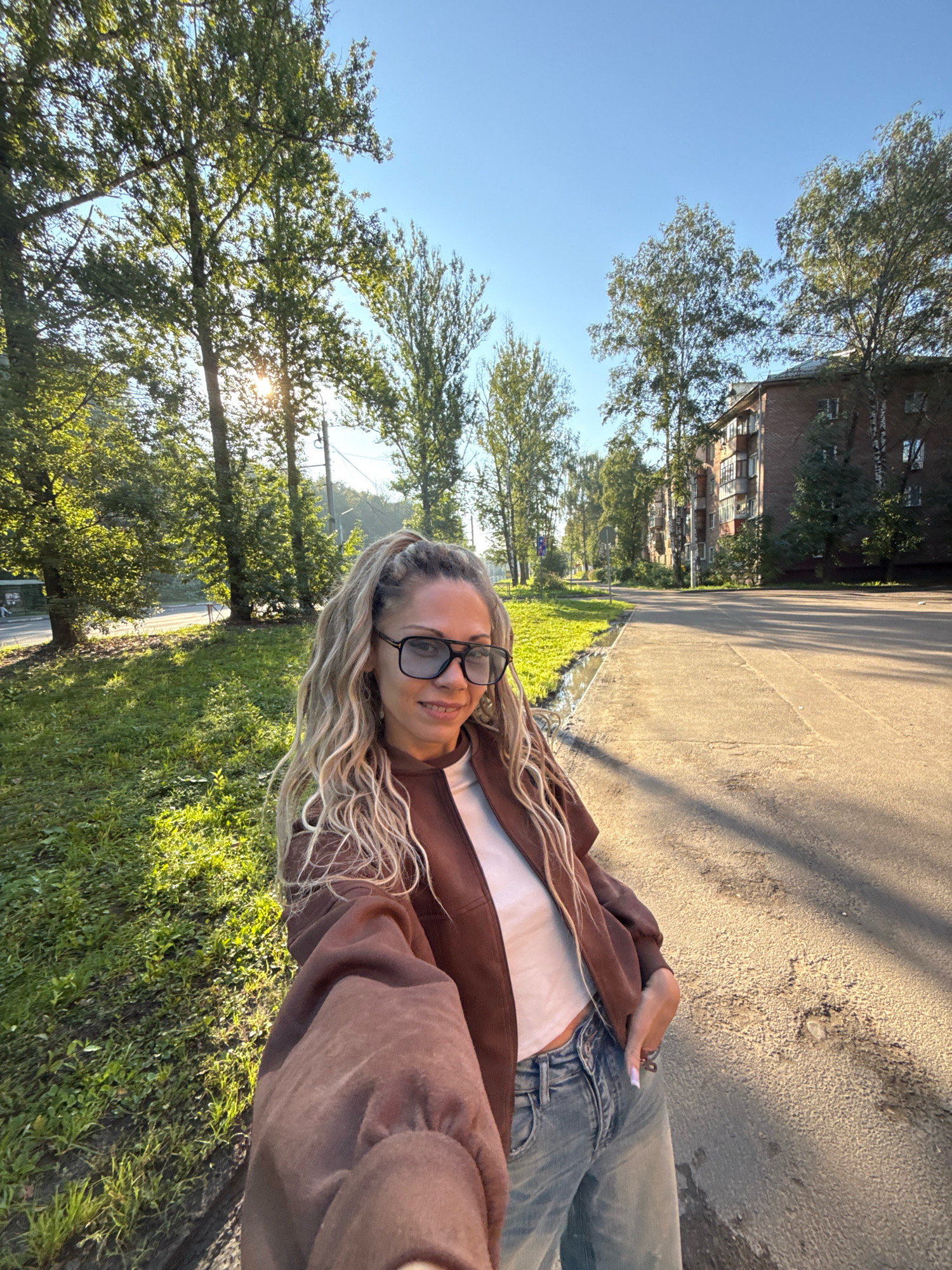 A woman with blonde, wavy hair pulled into a ponytail, wearing large dark-rimmed glasses, a white top, a brown jacket, and light blue jeans, is taking a selfie outdoors on a sunny day. She is standing on a sidewalk next to a paved road. To her left, there is a grassy area with trees. In the background, there are more trees and a multi-story brick apartment building. The scene appears to be a street in the city of Rusanovo, Russia.