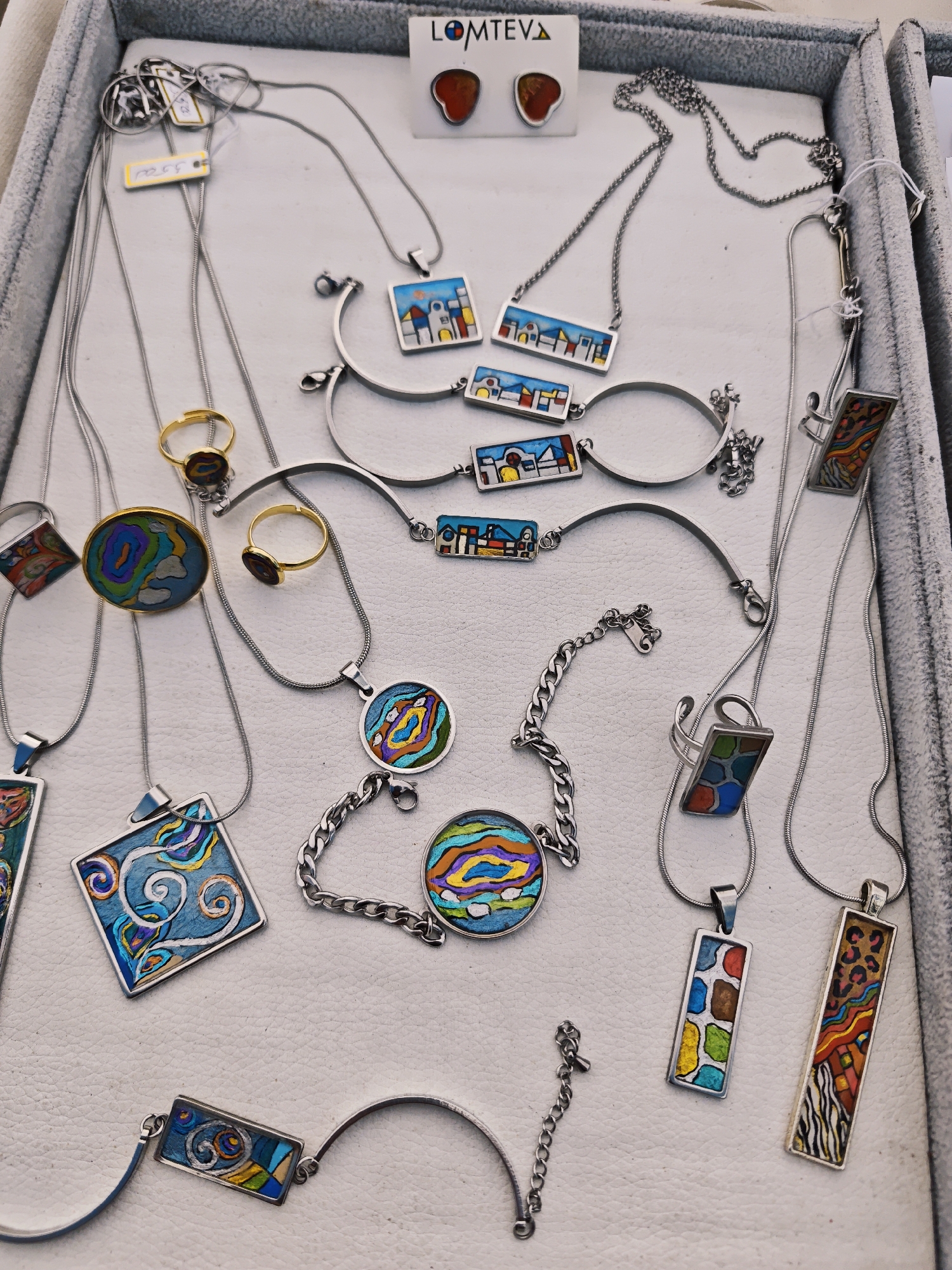 The image depicts a display of jewelry inside a grey tray. The jewelry includes necklaces, bracelets, rings, and earrings. The jewelry is silver with colorful designs. A small white tag with the word "LOMTEVA" is visible at the top. The scene is likely a jewelry display in Uglich, Russia.