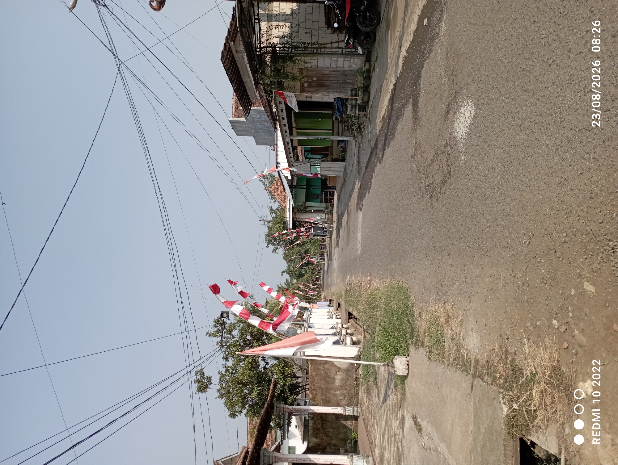 In Dukuhseti, Indonesia, a residential street is lined with Indonesian flags, indicating a national celebration. The flags, in red and white, are displayed on poles along the roadside and on top of some buildings. The street itself is paved with asphalt and appears to be narrow, with a sidewalk or drainage channel running alongside it. Several houses and small shops with tiled roofs and simple architectural styles line the street. Trees and vegetation are visible, suggesting a rural or semi-urban setting. There are also power lines stretching across the sky, typical of Indonesian villages. A red motorcycle is parked in front of one of the shops, and a white pillar stands by the entrance of one of the houses. The sky is clear and blue, suggesting a sunny day. The image was captured on August 23, 2026, at 08:26 AM, and is identified as being from a Redmi 10 2022 device.