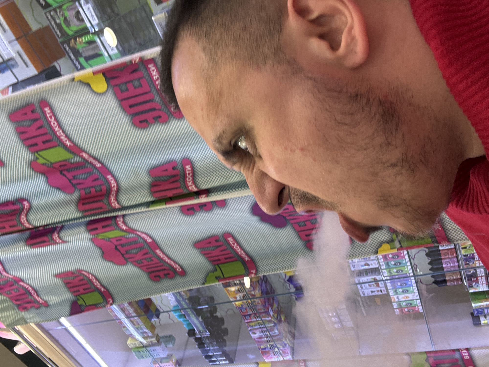 A man with short dark hair and stubble exhales vapor, possibly from an e-cigarette, in front of a display case containing various products, likely vaping supplies. The display case is marked with Russian text, partially visible as "ЭЛЕКТРОННАЯ ЖИДКОСТИ", and the products inside are likely arranged in rows and columns. The man is wearing a red garment. The location is Saint Petersburg, Russia, as suggested by the Russian text on the display case.