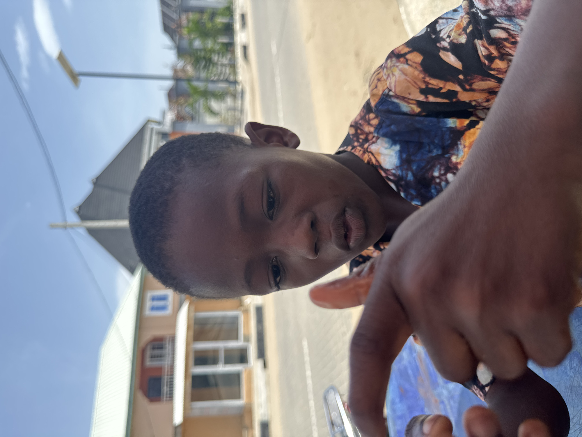 A close-up shot, slightly angled, reveals a young Black individual with short hair. They are wearing a patterned shirt in shades of blue, brown, and black. Their hand is prominently featured in the foreground.
In the blurred background, one can discern a street scene with buildings, including a two-story structure painted in hues of tan and green, with visible windows. A street light pole is also present, against a sky with scattered clouds.
Based on the visual cues of architectural style and general environment, the location is consistent with a setting in Badagry, Nigeria. No overt actions or interactions are discernible in the image.