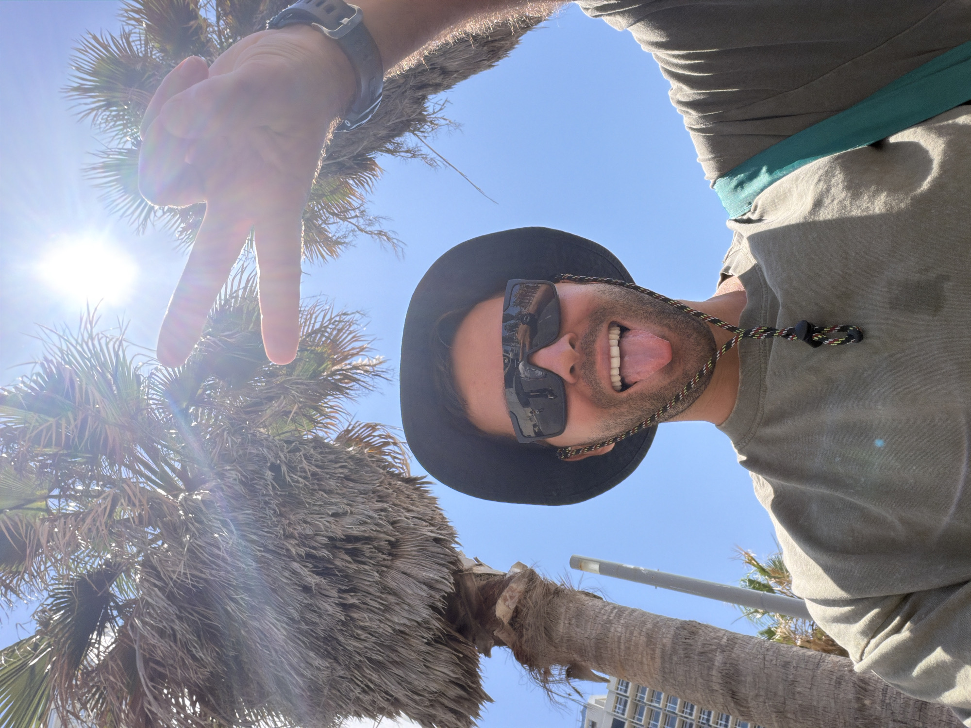 A man in a black bucket hat and sunglasses is sticking his tongue out at the camera. He is wearing a green t-shirt and has a teal bag strap over his shoulder. His right hand is raised, and he is making a peace sign. Behind him, two palm trees are visible, and the sun is shining brightly in the sky. The background also shows a glimpse of a tall, modern building, suggesting an urban coastal location in Tel Aviv, Israel. The lighting indicates it is daytime, likely late morning or early afternoon.