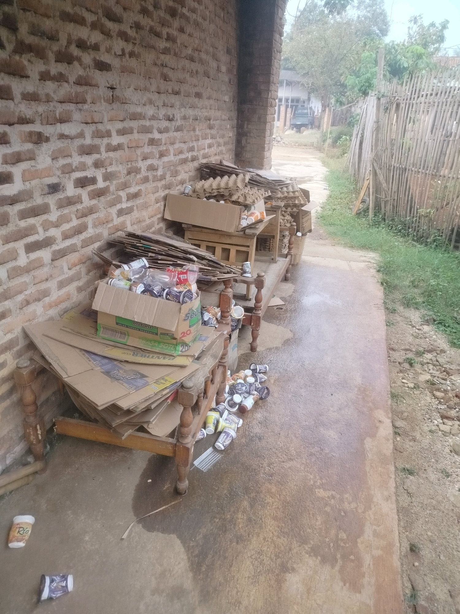 In Pandansari, Indonesia, an outdoor scene depicts a collection of recycled materials, primarily cardboard and aluminum cans, piled against a brick wall. A wooden bench or makeshift table holds a cardboard box filled with cans and plastic bottles, and additional cardboard is stacked on top. More cardboard and scattered cans are on the ground and under the bench. The environment suggests a residential or semi-rural area, with a dirt path, overgrown vegetation, and a bamboo fence visible in the background, alongside some structures and trees. The overall impression is one of a storage area for recyclables, possibly awaiting collection.