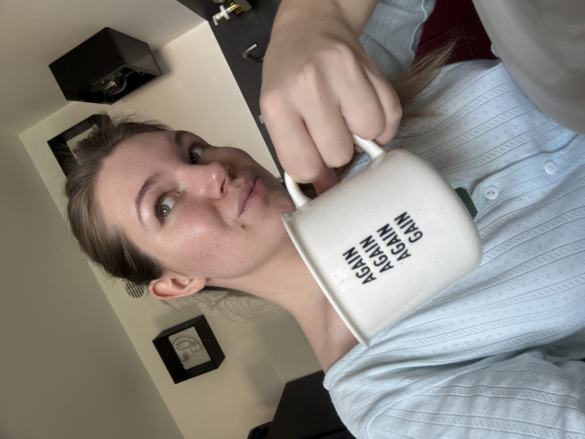 A close-up, high-angle indoor shot depicts a young adult female holding a white ceramic mug in her right hand, situated within a setting identified as Vozdovac Urban Municipality, Serbia. The female subject, possessing light skin and light brown hair pulled back, directs her gaze upwards and slightly to her right, exhibiting a subtle smirk. She is attired in a light blue, long-sleeved top featuring a fine eyelet knit pattern and white buttons, worn over a dark green garment. The white ceramic mug displays black text arranged vertically, stating "AGAIN AGAIN AGAIN GAIN," and incorporates a subtle green band at its base. The background to the left features a light-colored wall adorned with a black square shadow box frame enclosing minimalist line art of a face. A dark object, possibly a cabinet or appliance, is partially visible below this frame. A ventilation grate is present on the upper wall section. The background to the right reveals a dark grey or black countertop equipped with a metallic faucet and a clear pump bottle containing a yellow liquid, indicative of a soap dispenser. Another black square shelf or framed object, containing glass items, is partially visible above the countertop. The scene is brightly illuminated.