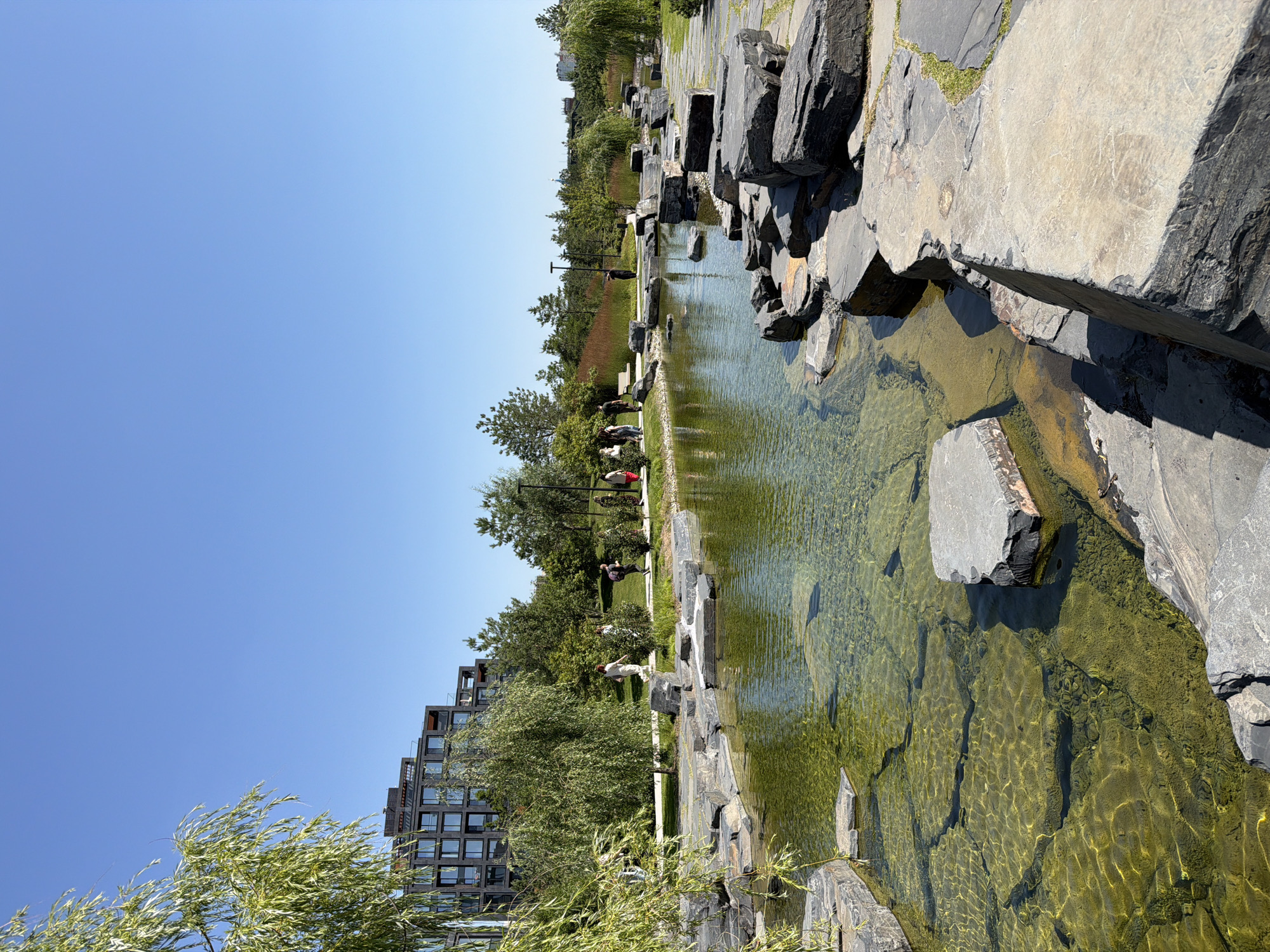 The image depicts a clear, sunny day in a modern park. In the background, a multi-story building with a dark facade and numerous windows dominates the skyline. In the mid-ground, a serene body of water, appearing as a pond or small lake, is surrounded by carefully placed large rocks of varying sizes and shades of grey and black. The water is remarkably clear, allowing visibility of the rocky bottom and some aquatic plants. A paved pathway meanders along the edge of the water and through the park, lined with grass and trees. Several individuals are observed walking on this pathway, engaged in casual strolls. A weeping willow tree with its characteristic long, trailing branches adds a touch of natural grace to the left side of the foreground, partially framing the scene. The overall environment suggests a well-maintained urban park designed for relaxation and recreation. The specific location is the Zaryadye Park in Moscow, Russia, known for its contemporary landscape architecture and proximity to the Kremlin.