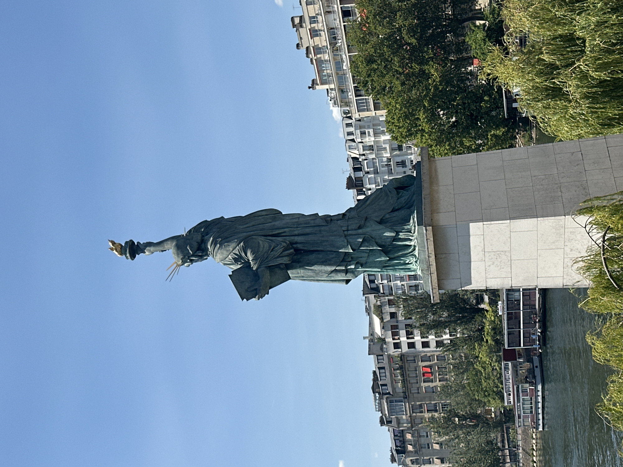 The image shows a full-body, rear-facing view of the Statue of Liberty replica in Paris, France, on a clear, sunny day. The statue stands on a stone pedestal by the Seine River. In the background, a row of multistory buildings lines the riverbank, with lush green trees interspersed among them. Two boats are docked on the river in front of the buildings. The statue is made of a green, weathered material, and it holds a book in its left arm and a torch, topped with a golden flame, in its raised right hand. The statue's crown has seven spikes, representing the seven continents and seven seas.