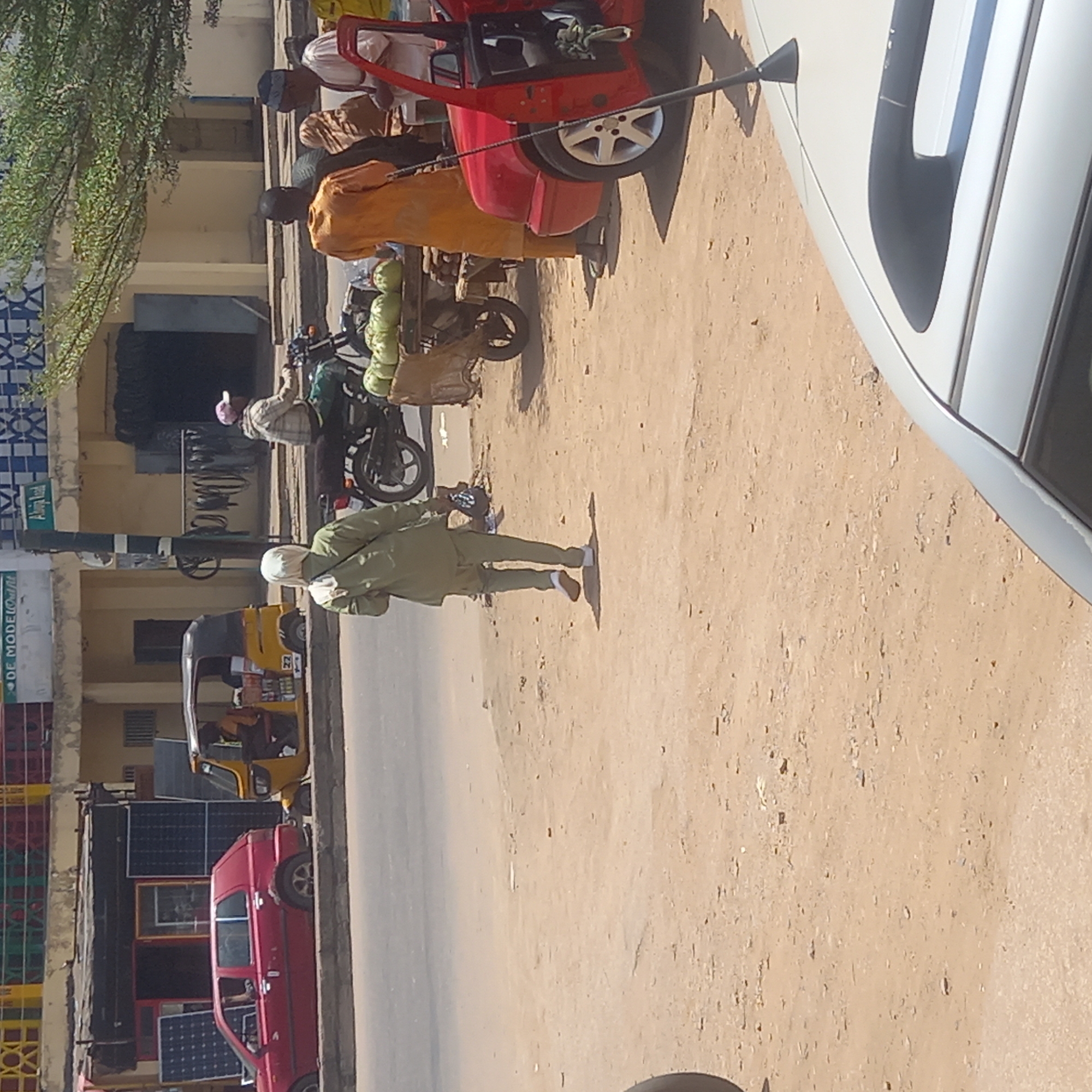 The image depicts a street scene in Dadi Riba, Nigeria. A man in a light green outfit is walking on the sandy ground next to a road. Several vehicles are parked on the side of the road, including a red three-wheeled vehicle, a yellow three-wheeled vehicle, a red car, and a white car partially visible on the right side of the image. People are standing near the parked vehicles. A motorcycle carrying what appear to be produce is also visible. Solar panels are visible on the side of a building along the road.