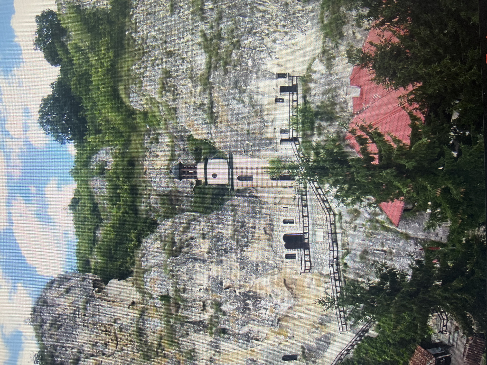 The image captures a historic monastery complex carved into and built upon a rugged, rocky cliffside under a partly cloudy blue sky. Lush green trees and vegetation cling to the cliffs and surround the structures.
The central feature is a stone bell tower with a conical roof, topped with a cross. It displays arched and circular windows. Several other stone buildings are integrated into the cliff face, featuring rectangular and arched window and door openings. Dark staircases or pathways with railings snake along the cliff, connecting various levels of the complex. To the lower right, partially obscured by dense green foliage, is a building with a prominent red-tiled roof.
The scene depicts a peaceful, static landscape with no people or activities visible. The clear sky and natural lighting suggest it is daytime, likely morning or afternoon, with good weather. The architecture and natural setting evoke a sense of history and spiritual tranquility. While some text appears on plaques above doorways, it is too indistinct to be read. The location context indicates this is a site near Varna, Bulgaria, consistent with such cliff monasteries.