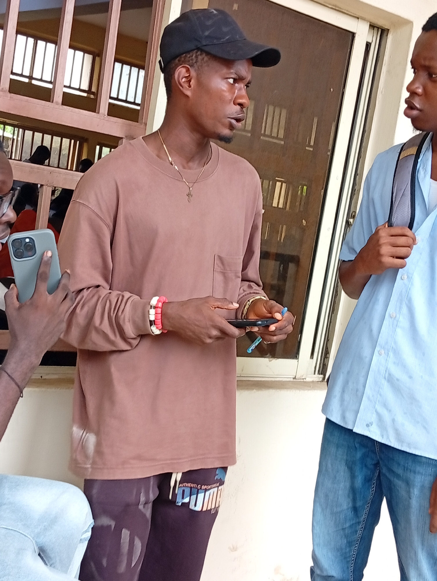 Three young men are standing outside a building with barred windows, likely in Ukukwa, Nigeria. One man, wearing a brown long-sleeved shirt and dark pants with a Puma logo, is holding a light blue phone in his left hand, looking intently at it. He has a gold chain with a cross pendant and two bracelets, one white and red and the other white, on his right wrist. Another man, wearing a light blue collared shirt and blue jeans, stands to his right, with a backpack slung over his shoulders. He is looking towards the first man, his mouth slightly open as if speaking. A third person, only partially visible on the far left, is wearing glasses and a light blue shirt, and is holding a grey phone, possibly taking a picture of the man with the brown shirt. The environment appears to be an outdoor area with a white wall and window frames.