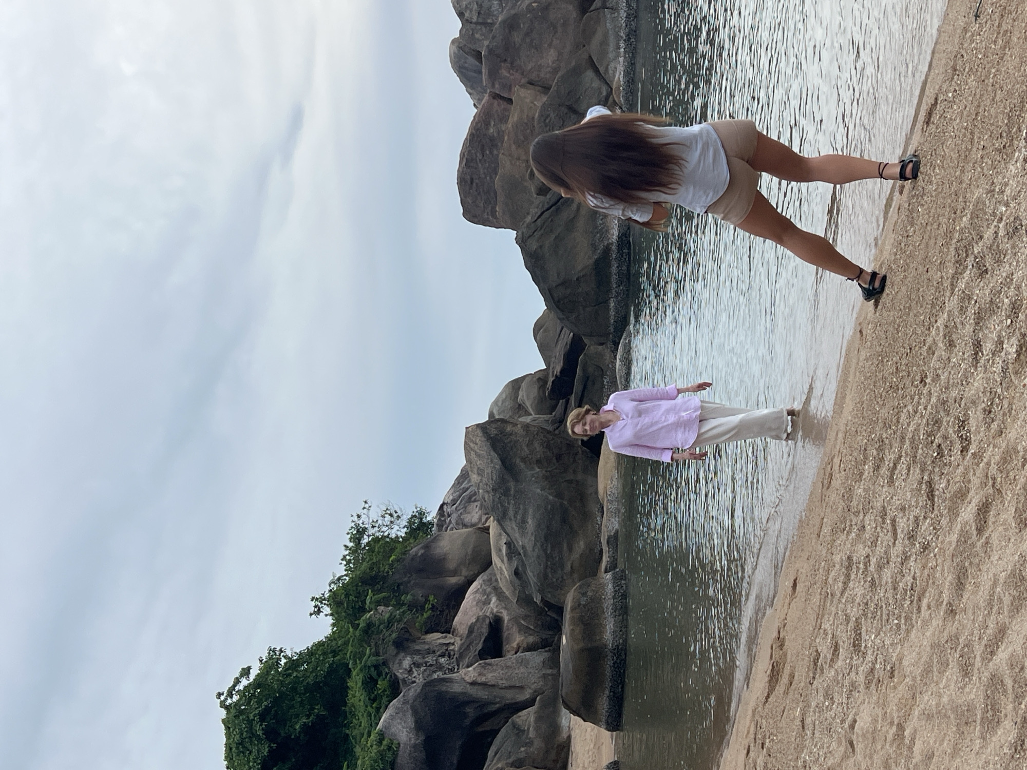 Two women are on a sandy beach in Baan Chaweng Noi, Thailand. One woman, wearing a light pink button-down shirt and white pants, stands near the water's edge, facing left. The other woman, with long brown hair and wearing a white shirt and beige shorts, stands on the sand, facing away from the viewer and holding a phone up as if taking a picture. She is positioned to the right of the first woman and slightly behind her. The background features large, dark, rounded rocks covered in green foliage, and the ocean with rippling water. The sky is overcast with pale clouds.