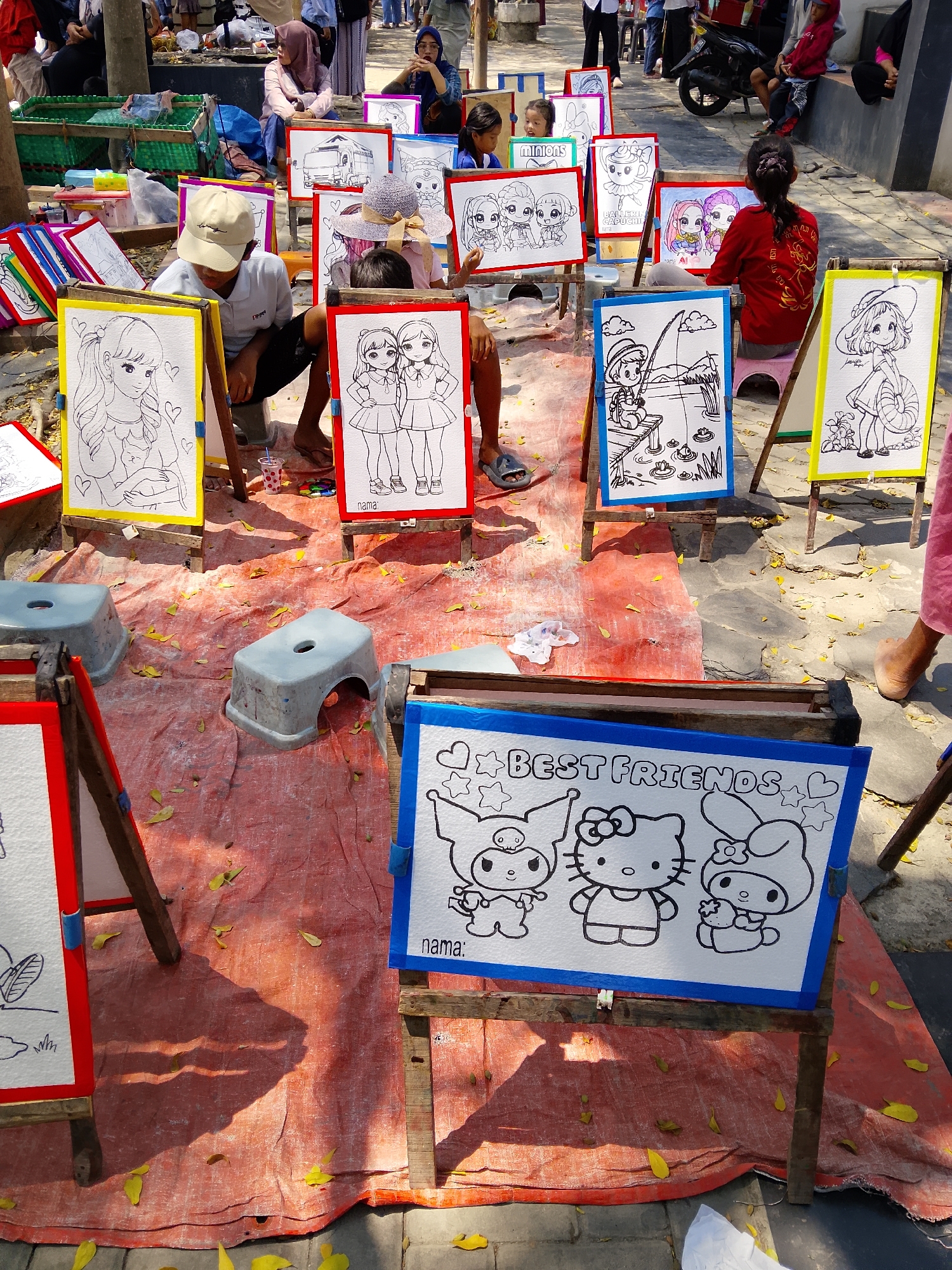 The image depicts an outdoor scene in Balun, Indonesia, where children are engaged in drawing. Several easels display various black and white drawings, primarily of cartoon characters and children. In the foreground, a white board with a blue border features a drawing of Hello Kitty and other Sanrio characters, with the text "BEST FRIENDS" at the top and "nama:" (which translates to "name:") below. To the left, another easel holds a drawing of a girl with long hair. In the middle ground, a boy wearing a beige cap and white shirt is seated, looking down at a drawing on a yellow-bordered easel. A red-bordered easel to his right displays a drawing of two girls. Further back, other children and adults are visible, some observing the drawings or engaged in other activities. The ground is covered with an orange tarp and scattered yellow leaves, suggesting an outdoor market or public space. Two small gray stools are placed on the tarp. The background shows a pathway, trees, and more people, indicating a bustling environment.