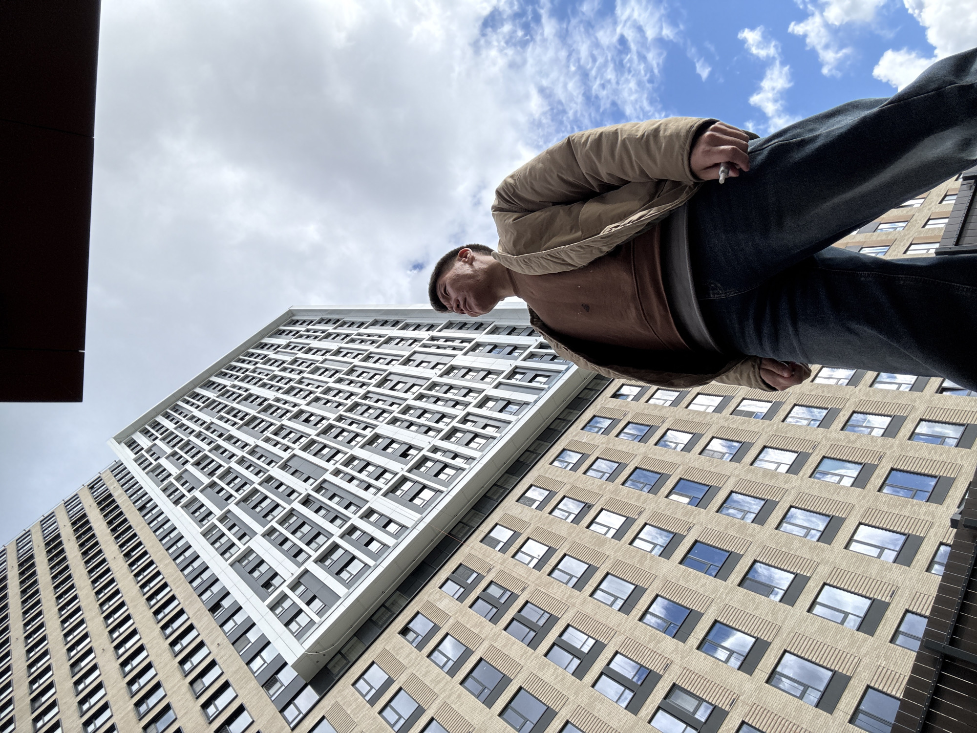A low-angle shot depicts a person standing between two buildings under a partly cloudy sky. The person is wearing a brown shirt, a tan jacket, and dark jeans, and is holding what appears to be a cigarette in their hand. The building on the left is tall and primarily white with a geometric pattern of windows. The building on the right is beige with regularly spaced windows. The buildings suggest an urban setting. The presence of the buildings and the person suggests an interaction with the urban environment. The specific location in Khimki, Russia, is not discernible from the image alone.