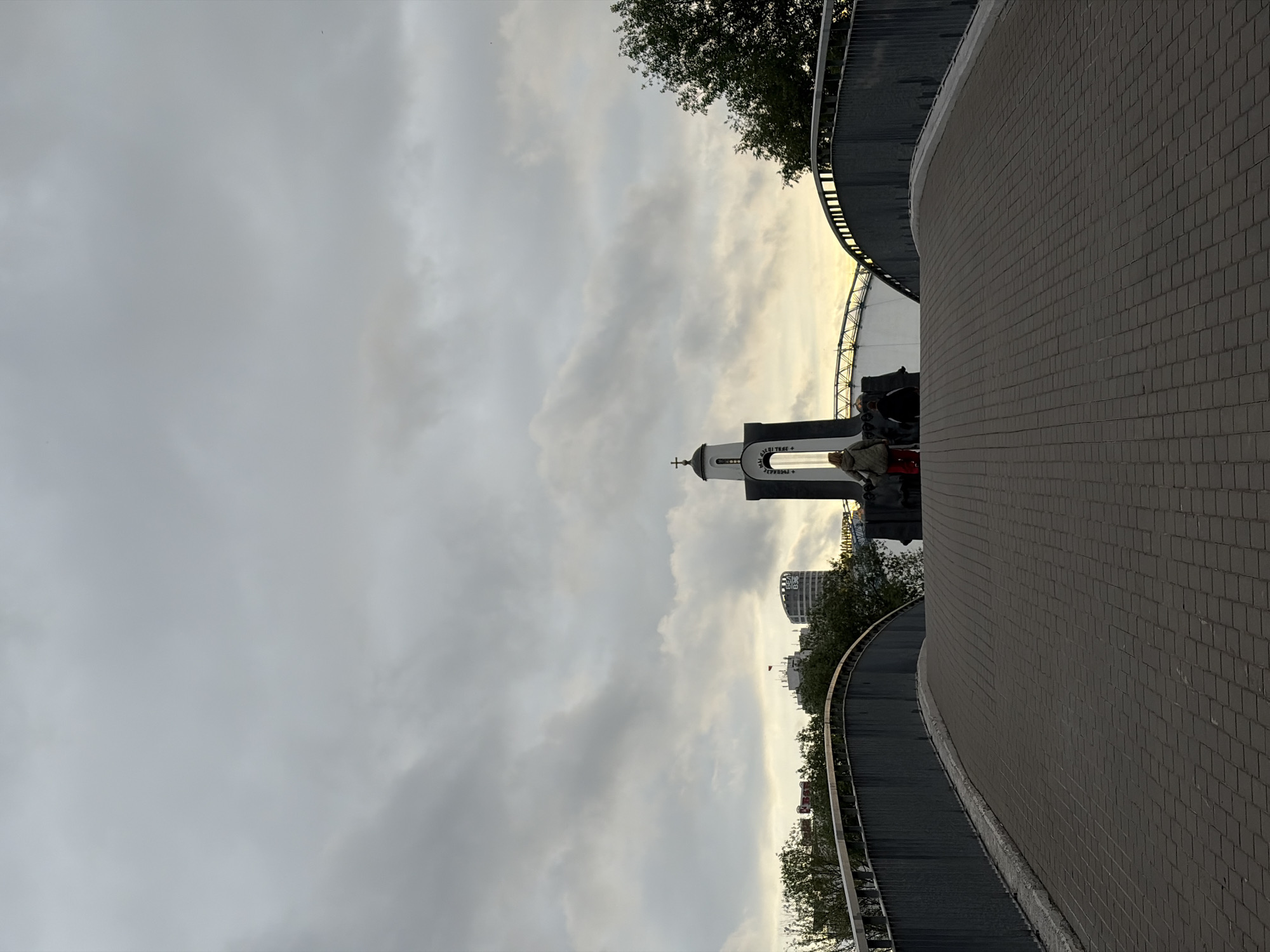 A low-angle, full shot captures a paved pedestrian bridge in Minsk, Belarus, leading towards a bell tower topped with a cross. The sky above is overcast with gray clouds, but a sliver of yellow light breaks through on the horizon, suggesting dawn or dusk. Two individuals, one wearing a beige jacket and red pants, and another in a dark jacket, stand near the bell tower. Behind them, modern buildings, including one with a curved facade, rise above a line of trees. A stadium structure with a white roof is visible in the background, to the right of the bell tower. The bridge is lined with a dark metal railing.