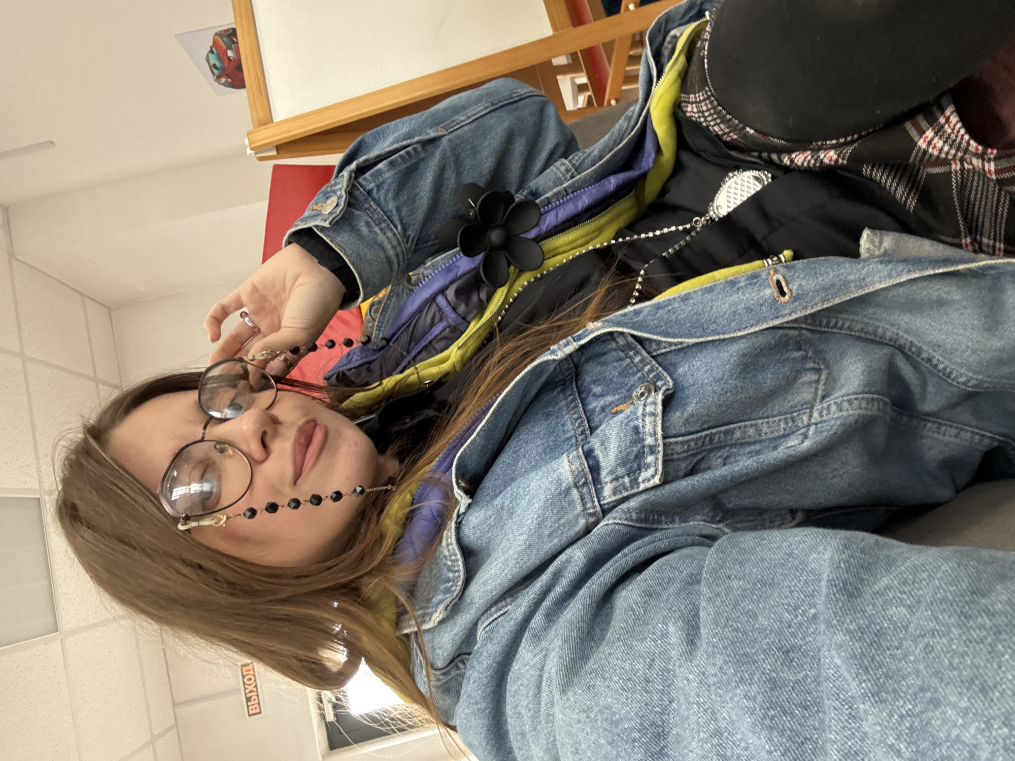 A young woman with long, brown hair is taking a selfie indoors, possibly in a classroom or office setting. She is wearing a blue denim jacket over a layered outfit that includes a black top, a yellow and purple scarf, and a black and red plaid skirt. She has on large, round glasses with a black beaded chain attached to one lens. A black flower-shaped clip is attached to her jacket. She is seated, with her right arm resting on her leg. The background is blurred but appears to be a brightly lit room with a white board and a sign that reads "ВЫХОД," which means "EXIT" in Russian.