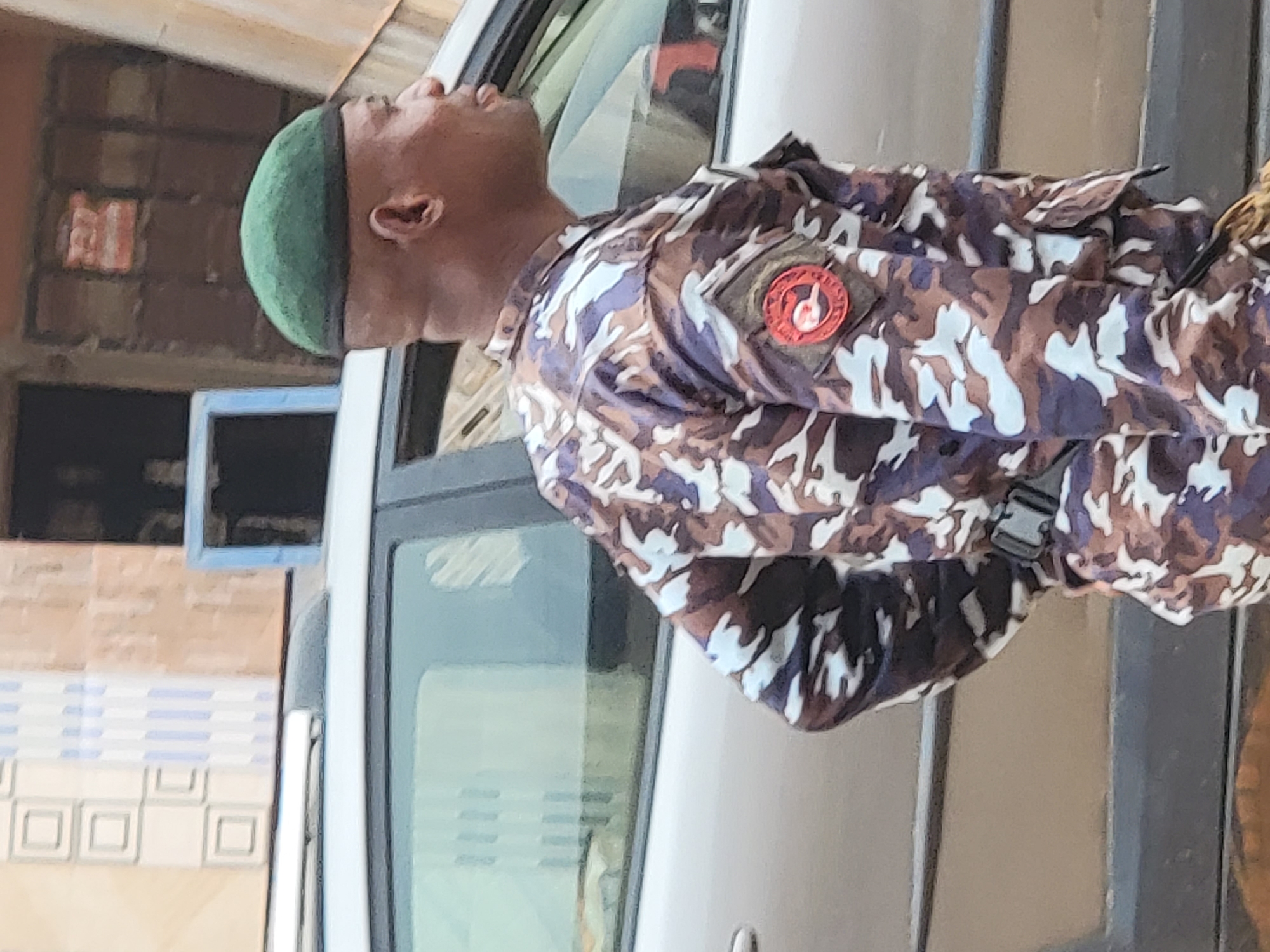 The image shows a male individual standing outdoors in Maiburiji, Nigeria, during the daytime. He is wearing a military-style camouflage uniform with a distinct pattern of brown, purple, and white. A plain green beret covers his head. On his left arm or shoulder, there is a circular red patch featuring a white or light-colored emblem. The specific details of the emblem are not clearly legible.
The person is positioned upright, appearing to look straight ahead or slightly upwards, with a composed expression. Behind him, a light-colored vehicle, possibly a van or large car, is partially visible. In the background, there is a building or wall adorned with patterned tiles, some of which display square designs. The bright, clear lighting indicates it is daytime, and there are no signs of adverse weather conditions. The uniform suggests the individual is security personnel or a member of an official service. No discernible text is visible on the uniform patch or elsewhere in the image.