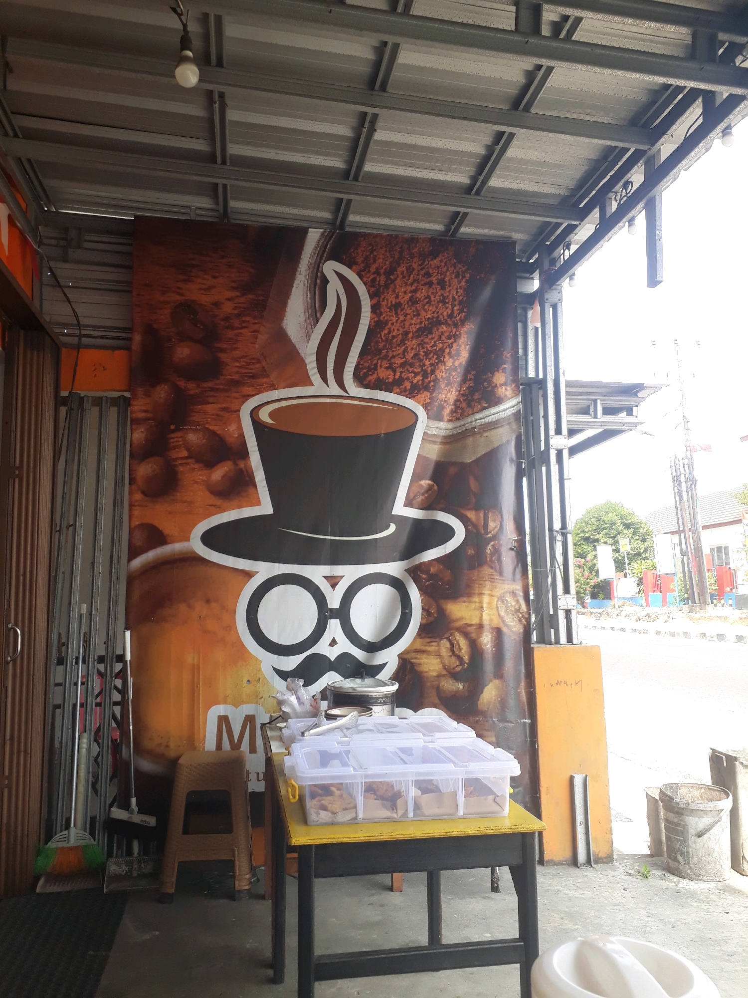 The image depicts the facade of a coffee shop or eatery in Pangkalpinang, Indonesia. The main focus is a large banner featuring a stylized illustration of a coffee cup with a hat and glasses, set against a background of coffee beans and ground coffee. To the left, a brown accordion-style gate and metal poles are visible, suggesting the entrance to the establishment. In front of the banner, a yellow table holds a transparent plastic container filled with food items and a pair of metal tongs. A small brown plastic stool is placed to the left of the table. To the far left, a broom with green and orange bristles leans against the wall. The scene is covered by a corrugated metal roof supported by metal beams. In the background, a street with cars and buildings can be seen, along with trees and utility poles. The overall setting appears to be an open-air seating area or storefront.