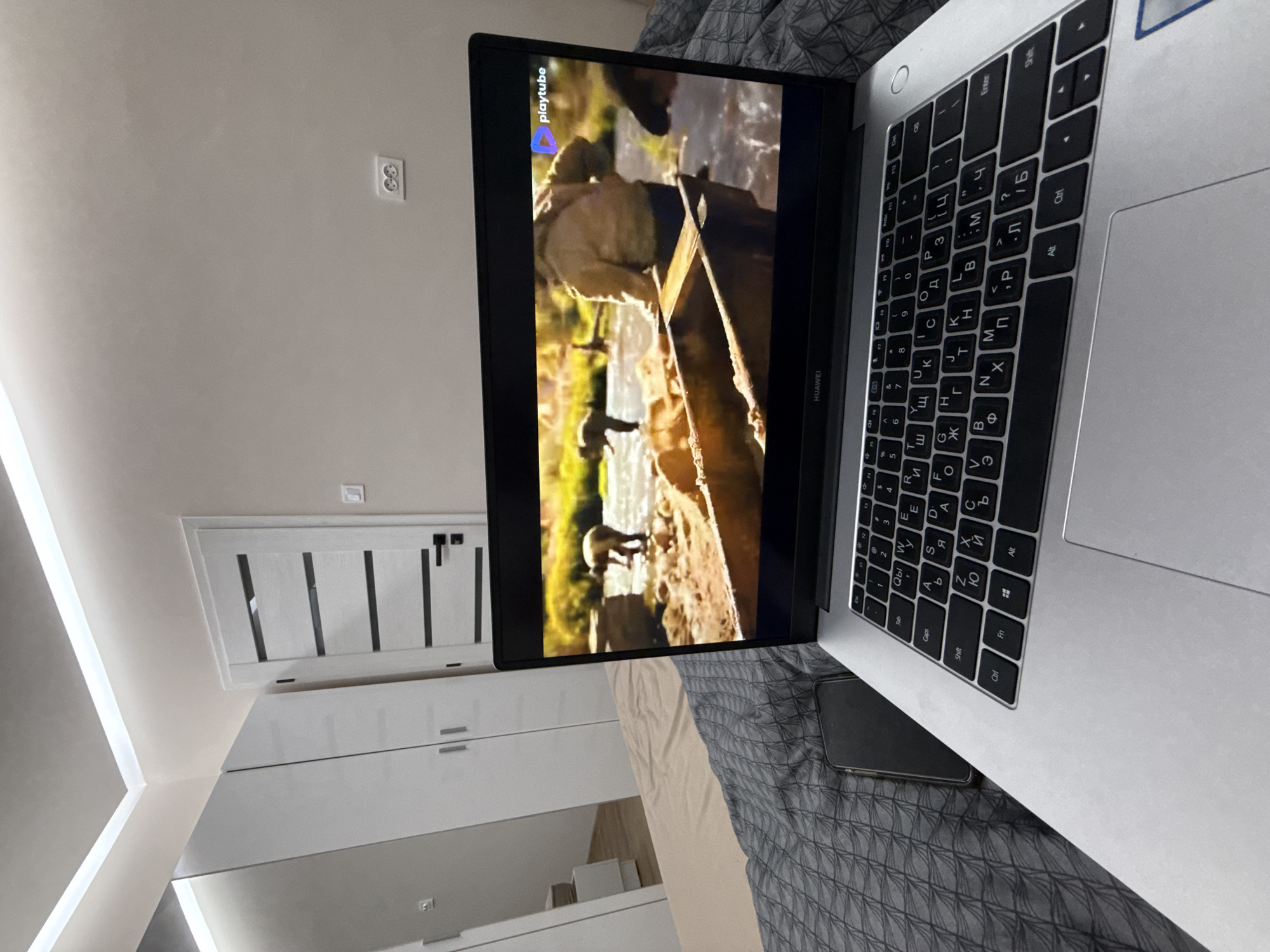 A close-up indoor shot shows a laptop displaying a YouTube video, positioned on a bed with a patterned grey duvet. A black smartphone rests beside the laptop. The room's interior is visible in the background, featuring white walls, an electrical outlet, a door with vertical black accents, and a white storage unit. The focus is on the laptop and immediate surroundings.