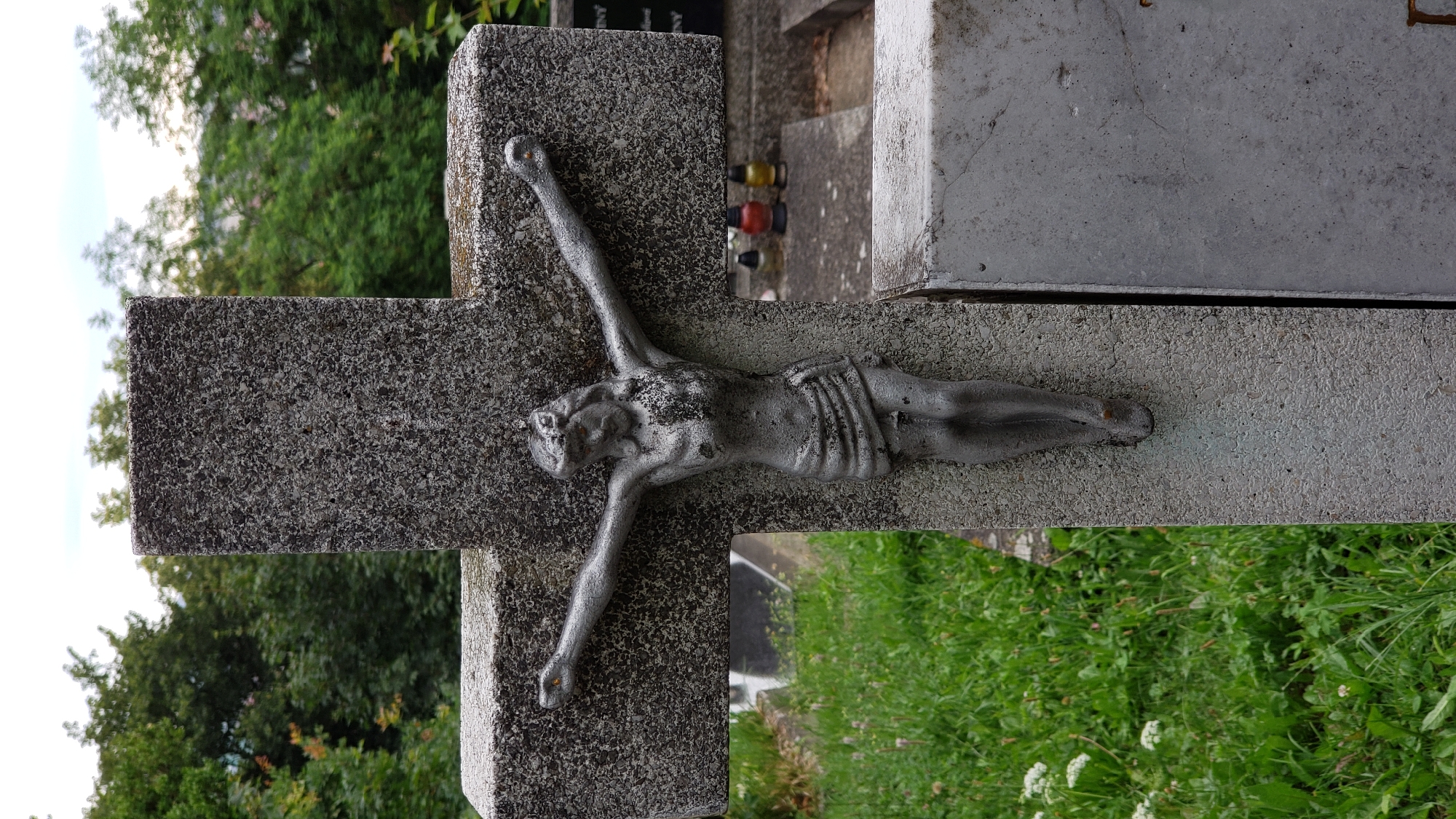 The image shows a close-up view of a gravestone in a cemetery. The gravestone is a large, gray concrete cross with a metallic sculpture of Jesus Christ crucified. The sculpture is detailed, showing Jesus with a crown of thorns and a loincloth. The cross is weathered, with some dark staining and a rough texture. To the left of the cross is a patch of green grass with some white wildflowers. In the background, there are trees, other gravestones, and a cloudy sky. The overall impression is one of solemnity and remembrance. This scene is located in the cemetery in Prešov, Slovakia.