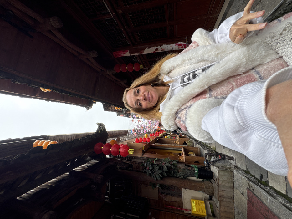 A selfie captures a light-skinned female subject in a narrow, stone-paved street within the city of Furong, China, under an overcast sky. The female, positioned in the foreground, has long, wavy blonde hair and a faint smile directed at the camera. She wears a white hooded sweatshirt with a dark graphic print under a pink, white, and grey plaid coat featuring extensive white faux fur trim on the hood, collar, and cuffs, and a fringed lower hem. Her right arm is extended, holding the camera, with her left hand partially visible.
The street is flanked by traditional multi-story wooden buildings characterized by dark timber structures and tiled roofs with distinctive upturned eaves. Red lanterns are prominently displayed, hanging in vertical arrangements from the building overhangs; stacks of three are visible on the left and four on the right. Further back, a dense canopy of small, colorful decorative elements is strung across the alleyway. Orange, spherical ornaments are also visible along the edges of the tiled roofs. On the left side of the street, a shopfront displays various items on exterior wooden shelving, alongside a green potted plant and a yellow utility box. The building on the right features closed dark wooden doors or panels.