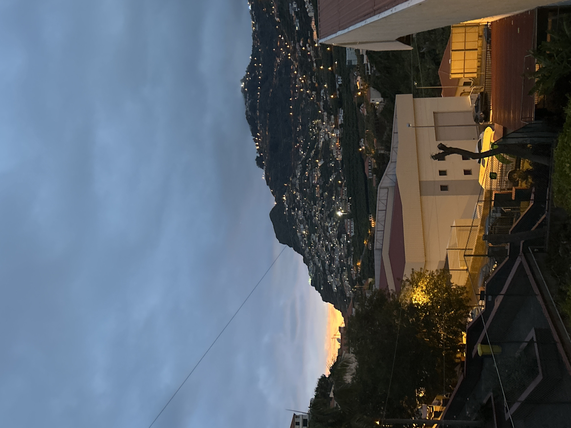 The image shows a view in the city of Câmara de Lobos, Portugal. A hillside dotted with lights is visible under a cloudy sky. The sky transitions from a light blue-gray to a golden hue near the horizon. In the foreground, buildings are illuminated, indicating it is evening. There is a tree and a fence visible in front of the buildings.