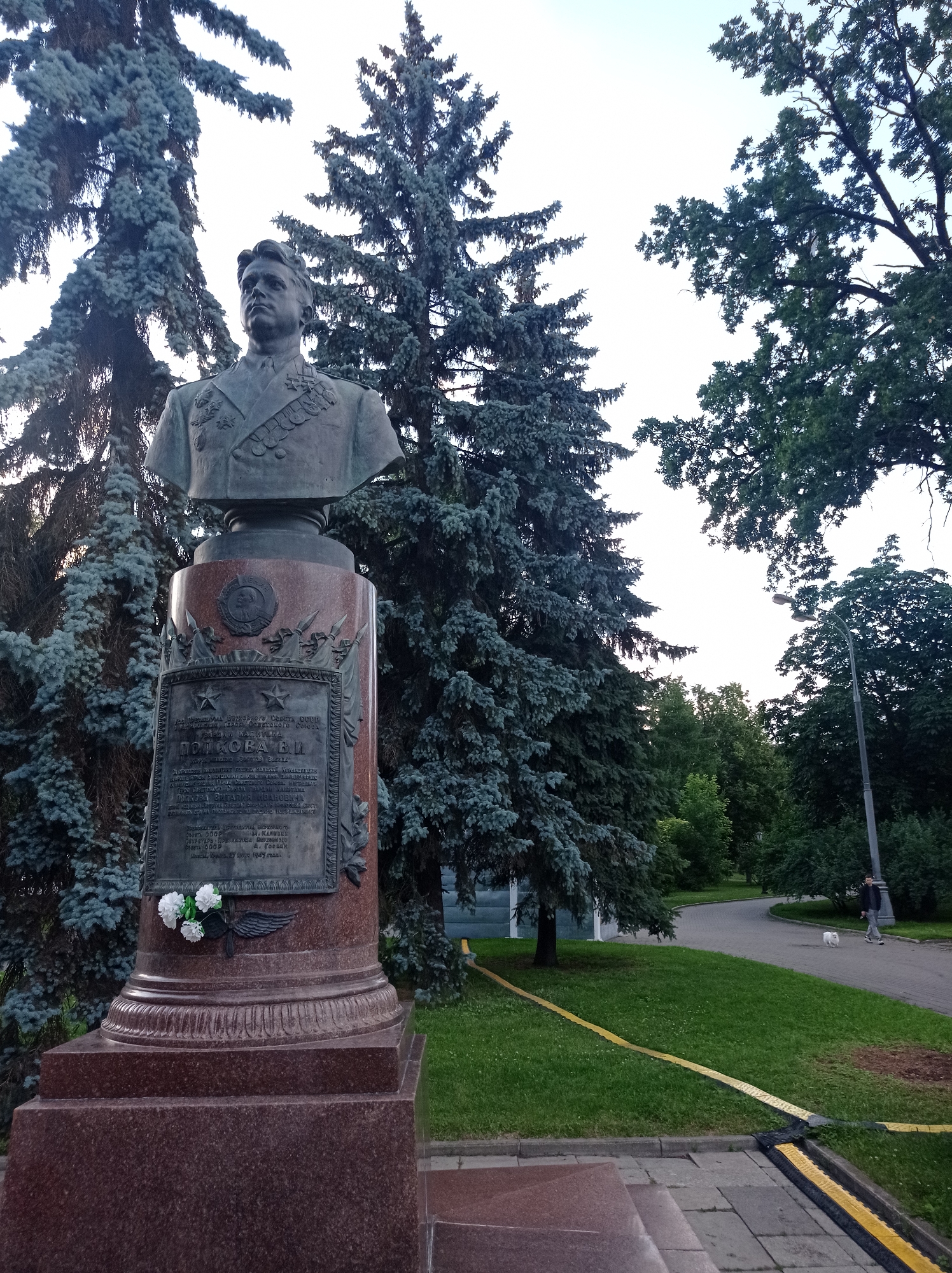 A bronze bust of a man in military uniform with medals is mounted on a granite pedestal in a park. The bust depicts a man with short, dark hair, a serious expression, and a medal-laden jacket. The pedestal is inscribed with text and adorned with a winged emblem and a small bouquet of white flowers. Behind the monument, tall evergreen trees with blue-green needles frame the scene. In the background, a paved pathway winds through a grassy area with more trees. A lamppost is visible to the right, and a person is walking a small white dog in the distance. The overall setting appears to be a public park in Moscow, Russia.
