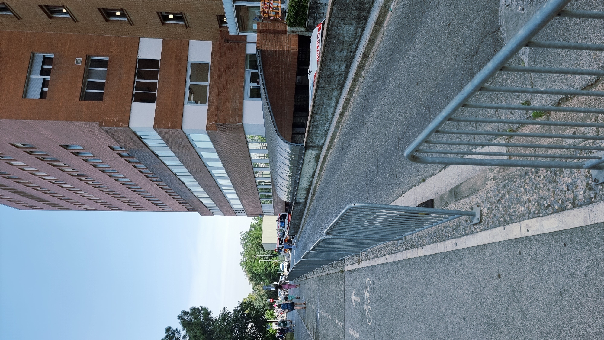 The image depicts a wide view of a street with a tall, modern brick building on the right and a sidewalk on the left. The building has many windows and appears to be an office building or hospital. A covered walkway with a translucent roof is visible in front of the building. On the sidewalk, several people are walking, and there are markings on the ground indicating a bike lane and pedestrian path. A metal fence runs along the edge of the sidewalk and road. Ambulances are parked on the road in front of the covered walkway. The scene appears to be a public area with traffic and pedestrian activity.