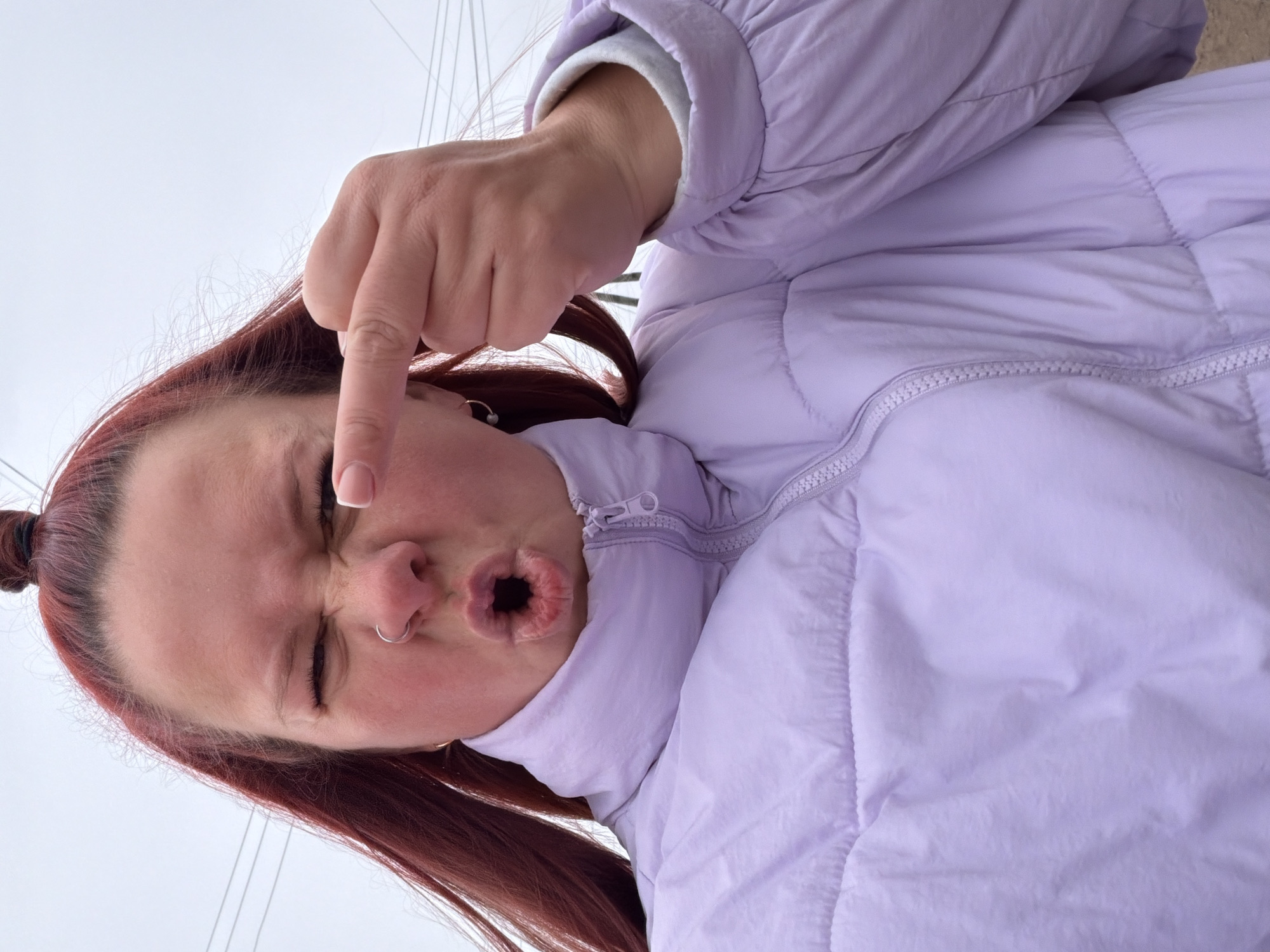 A woman with auburn hair pulled into a bun is looking down at the camera and gesturing with her middle finger pointing down. She is making a kissing face, with her lips pursed and round. She has a nose ring and is wearing a light purple puffy jacket. Overhead, there are power lines. The location is potentially Chaikovsky, Russia.