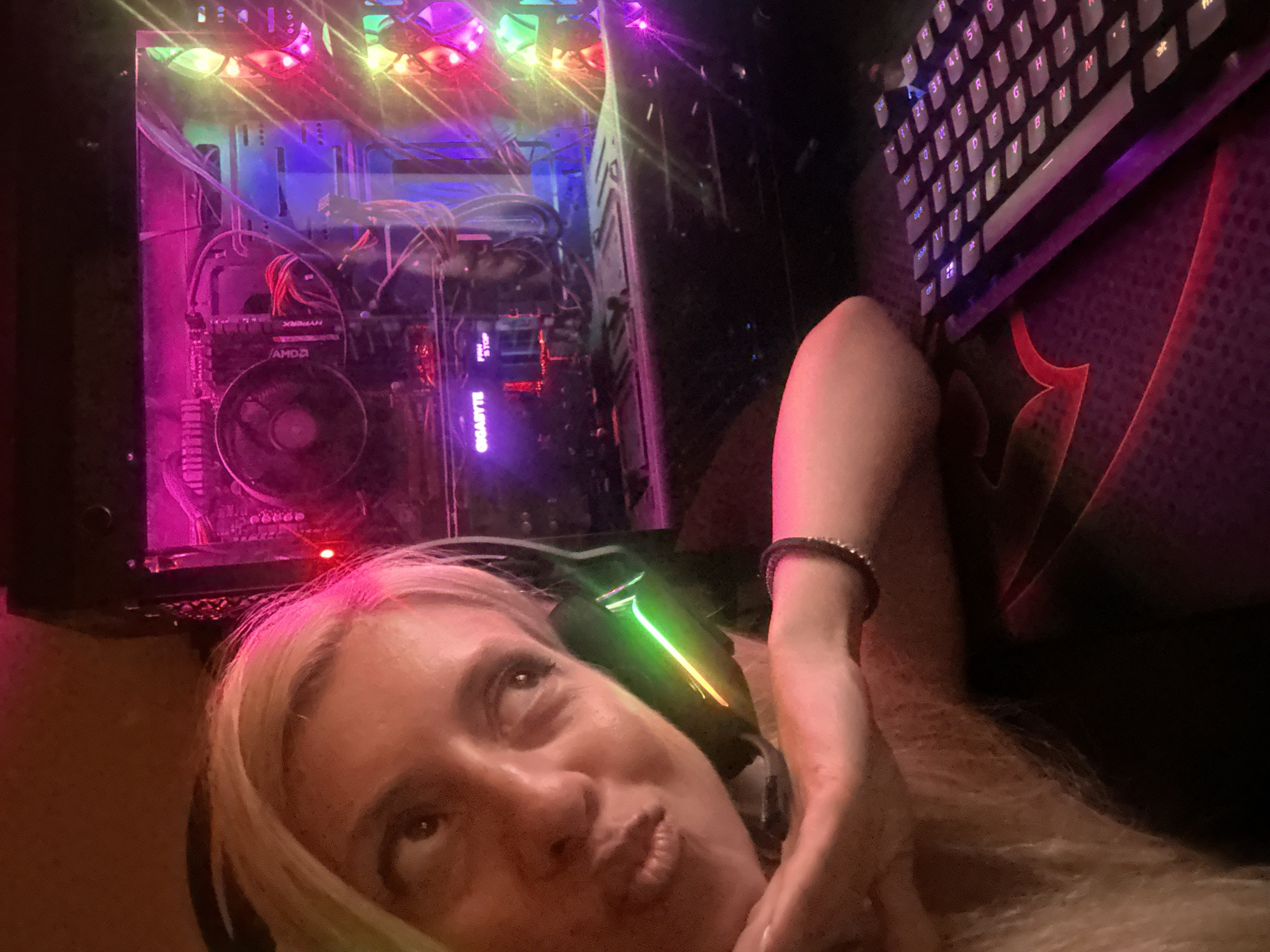 The image shows a close-up of a woman with blonde hair, wearing headphones, and making a kissy face. She is resting her chin on her hand. Behind her is a gaming computer with colorful LED lights. A keyboard is visible in the foreground. The scene appears to be indoors, likely in a home environment. There is no indication of the location being in Pazardzhik, Bulgaria, or any other specific city. The lighting is dim and atmospheric, with the LEDs from the computer and keyboard providing the primary illumination. The woman's expression suggests a playful or affectionate pose. The overall impression is of someone engaged in or enjoying a gaming session.