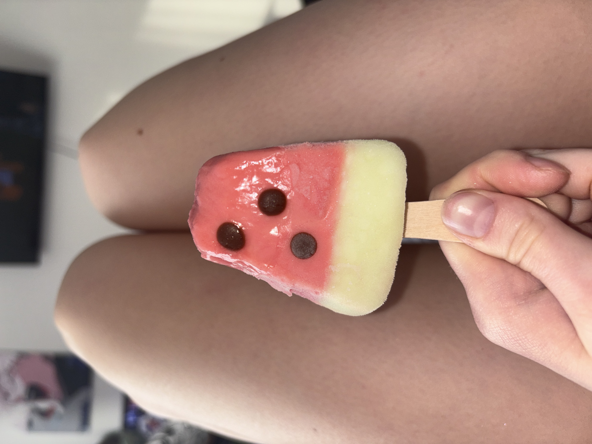 The image presents a close-up view of a hand holding a watermelon-shaped popsicle against a blurry background featuring a person's legs and indistinct indoor elements.
The main subject is a colorful popsicle, designed to resemble a slice of watermelon. Its upper portion is a vibrant pinkish-red, transitioning to a pale yellow or lime green at the bottom, mimicking the fruit's rind. Three dark brown, round chocolate chips are embedded in the pink section, serving as watermelon seeds. The popsicle appears slightly melted and frosty. It is held by a light-skinned hand, with visible fingernails, gripping a simple wooden stick.
In the soft-focus background, the smooth skin of a person's thighs or legs is prominently visible. A small, dark mole is noticeable on one of the legs. Further behind, very blurry shapes and light sources suggest an indoor setting, possibly including furniture or a screen. The overall lighting appears soft and consistent, indicating an indoor environment, perhaps under artificial light or diffused natural light. There is no visible text in the image.