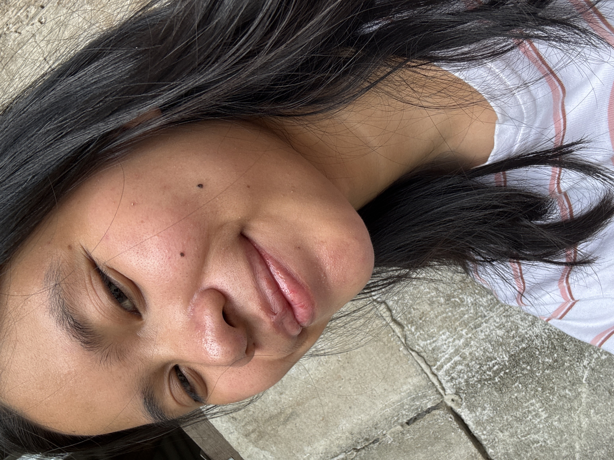 A close-up selfie of a young woman with long, dark hair is taken outdoors, against a weathered concrete wall. The woman is facing towards the camera, with her head tilted slightly to her left. Her eyes are partially closed, and she has a gentle smile on her face. Her skin appears to have some texture, with visible pores and a few small moles. She is wearing a white t-shirt with thin pink and brown stripes. The background consists of a rough, grey concrete surface that shows signs of age and weathering, with visible cracks and discolored patches. The lighting suggests it is daytime, with soft natural light illuminating the scene. The overall impression is a casual, candid portrait. The location is in the city of East Jakarta, Indonesia.