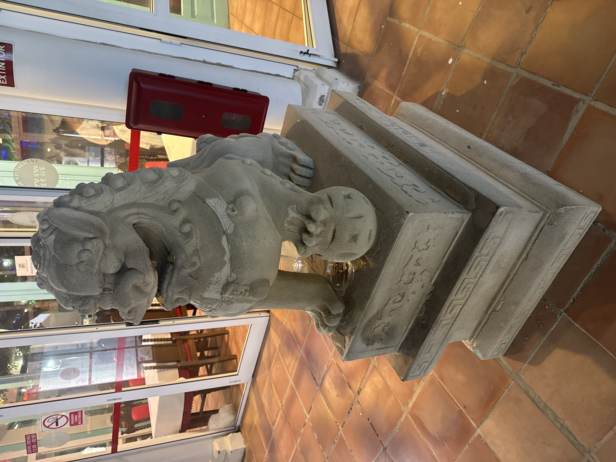The image shows a stone lion statue on a multi-tiered base, positioned outside a building. The building appears to be a restaurant, based on signage visible in the window. The signs read "FU EVI 富贵酒家" and "HORARIO-TIME 12:00-00:00", with a "PROHIBIDO FUMAR EN LA TERRAZA" (No Smoking) sign. A red fire extinguisher box is mounted on the wall near the building's entrance. The statue rests on a red-tiled surface. Given the restaurant’s name in Chinese, and the presence of the stone lion (a common Chinese architectural feature), it is likely that the image was taken in a Chinese restaurant located in Tías, Spain.