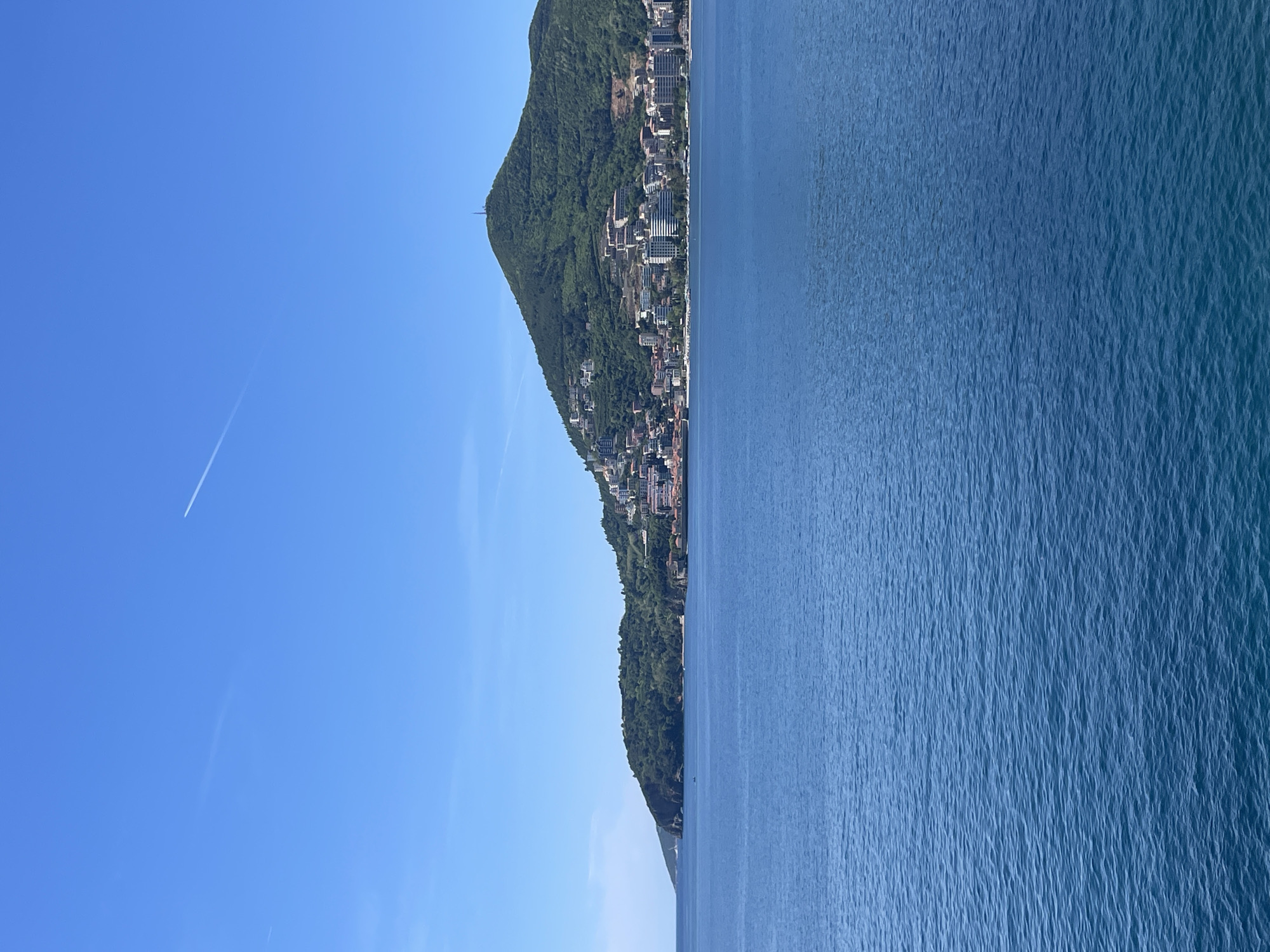 The image, when viewed in a corrected landscape orientation, presents a picturesque coastal scene, characteristic of Budva, Montenegro, under a brilliant blue sky. The right side of the frame is filled with the vast, calm waters of the sea, displaying various shades of blue with subtle ripples across its surface, suggesting a tranquil state.
On the left, a large, lush green mountain rises steeply from the water's edge. Its slopes are densely populated with numerous buildings, forming a bustling coastal town. These structures, ranging from multi-story modern buildings to smaller residences, are integrated into the verdant landscape and extend up the hillside. A slender, dark structure, possibly an antenna or tower, is faintly visible near the mountain's highest point.
The upper portion of the image is dominated by a wide expanse of clear, cloudless blue sky. A single white contrail from an aircraft diagonally streaks across the upper left section, indicating distant air traffic. The entire scene is bathed in bright, natural sunlight, suggesting it is daytime, likely morning or early afternoon. The weather is clear and sunny. While no specific human activity is discernible, the presence of the town implies a vibrant community. No visible text is present in the image.