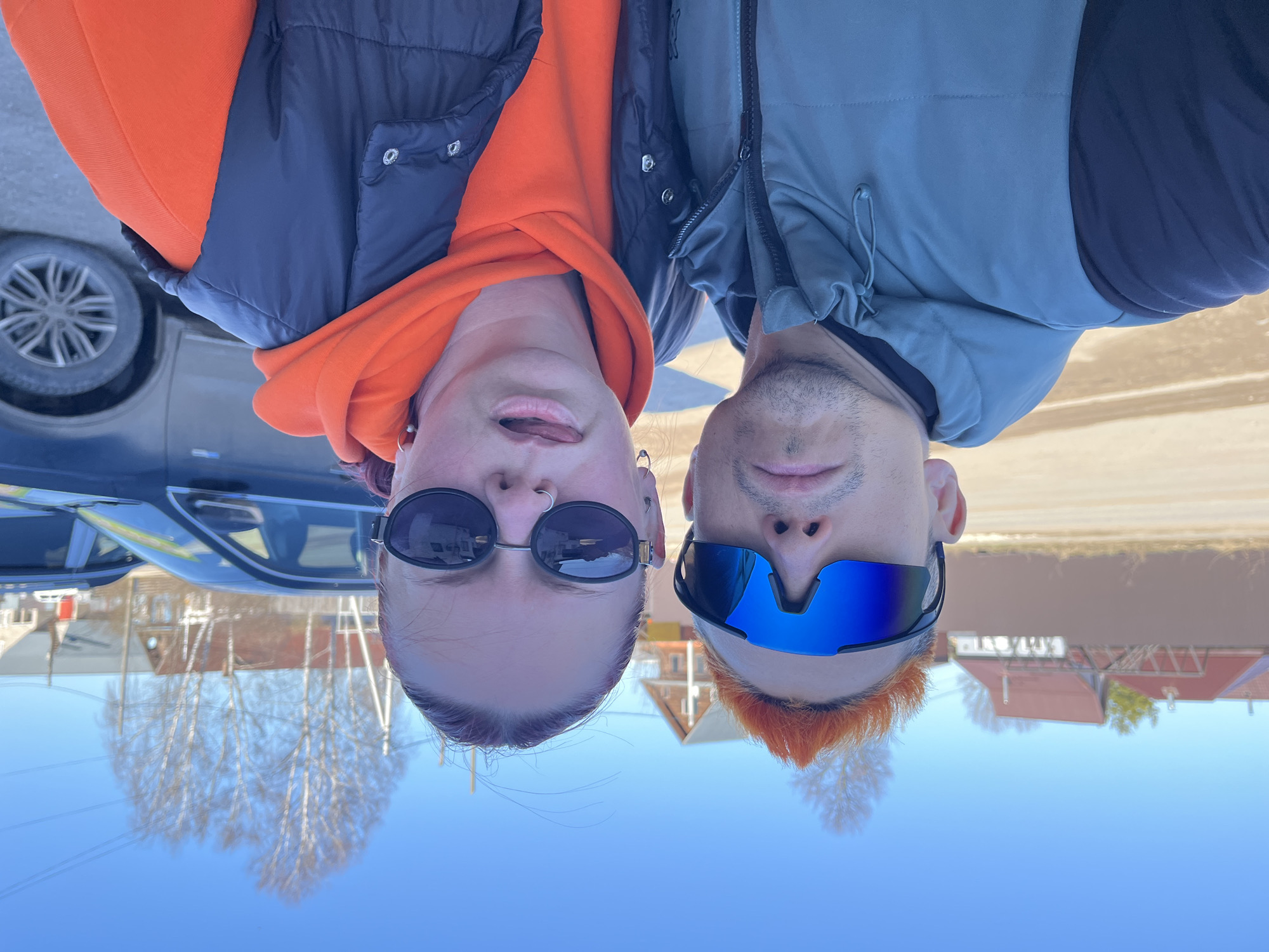 Two individuals, a woman and a man, are positioned upside down in the frame. The woman on the left is wearing sunglasses, an orange hoodie, and a black vest. The man on the right is wearing blue sunglasses and a light grey jacket. They are near a dark-colored car. The location is in the city of Chaikovsky, Russia, based on the surrounding environment, including the style of architecture visible in the background, such as houses with pitched roofs. A clear blue sky is visible. Bare trees are also in the background, reflecting off the car. The ground appears to be a mix of paved and unpaved surfaces.