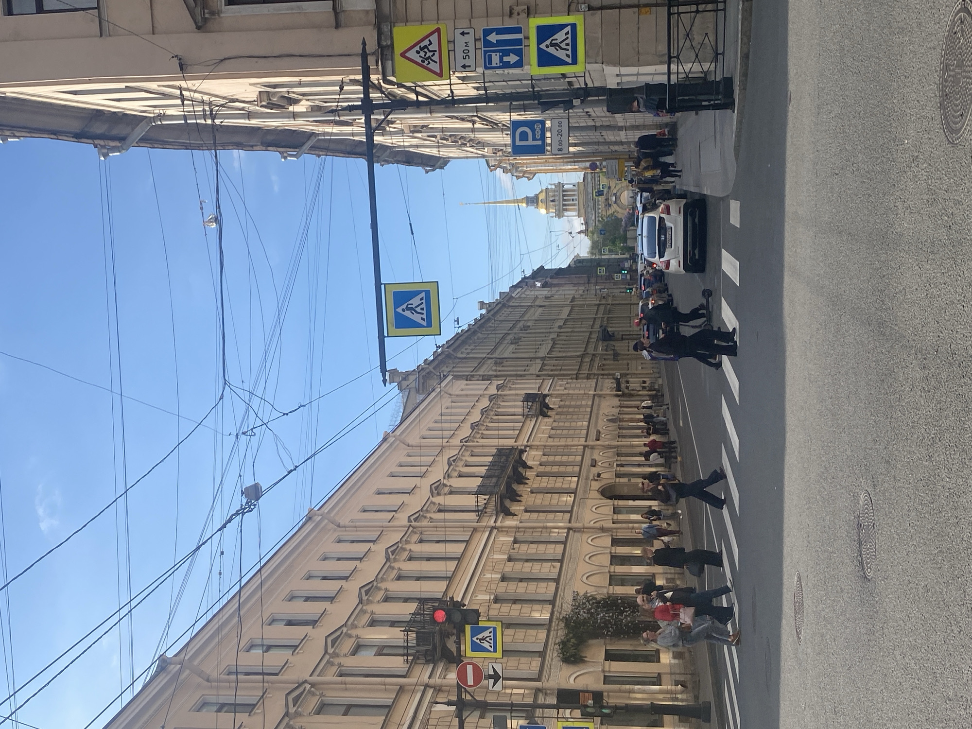 The image captures a street scene in Saint Petersburg, Russia. The view is along a straight road lined with multi-story buildings. Numerous overhead power lines are strung between the buildings. Several pedestrians are crossing the street at marked crosswalks. A white car is visible in the middle of the street along with a few other vehicles in the distance. Traffic and pedestrian signs are posted on poles alongside the road. The golden spire of a cathedral or other prominent building is visible in the distance.