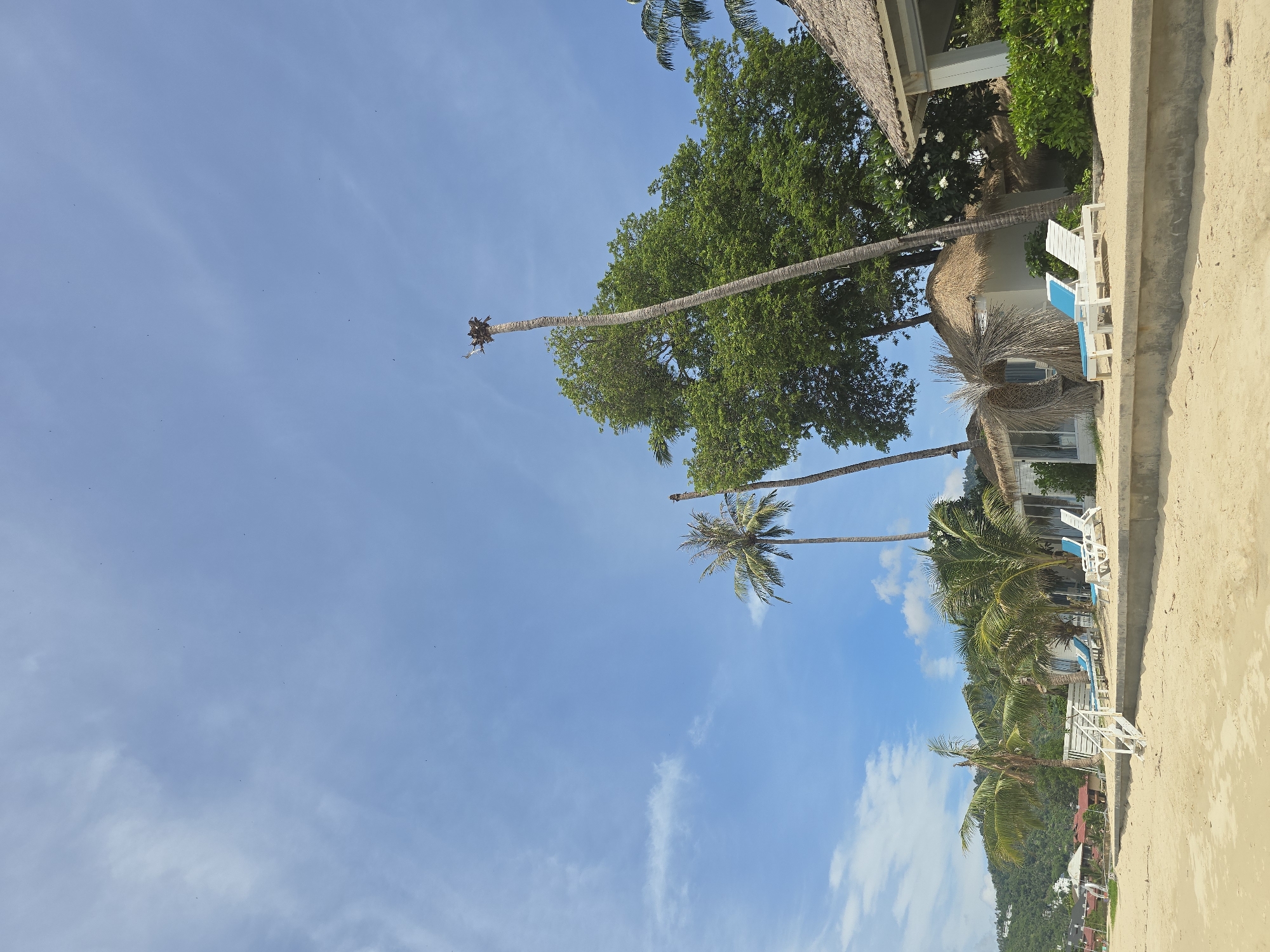 The image shows a beach scene in Baan Lamai, Thailand, under a bright blue sky with scattered white clouds. In the foreground, a sandy beach slopes down from a concrete retaining wall. A white wooden staircase with railings leads up from the beach to the retaining wall.
Along the retaining wall are several thatched-roof bungalows and one larger building with a light-colored roof. In front of the bungalows are lounge chairs, some white and some with blue cushions. Palm trees are scattered throughout the scene, with some tall, slender palm trees and one larger, fuller tree. In the background, a lush green hillside with buildings can be seen. The overall impression is a tropical resort setting. There are no people visible in the image.
