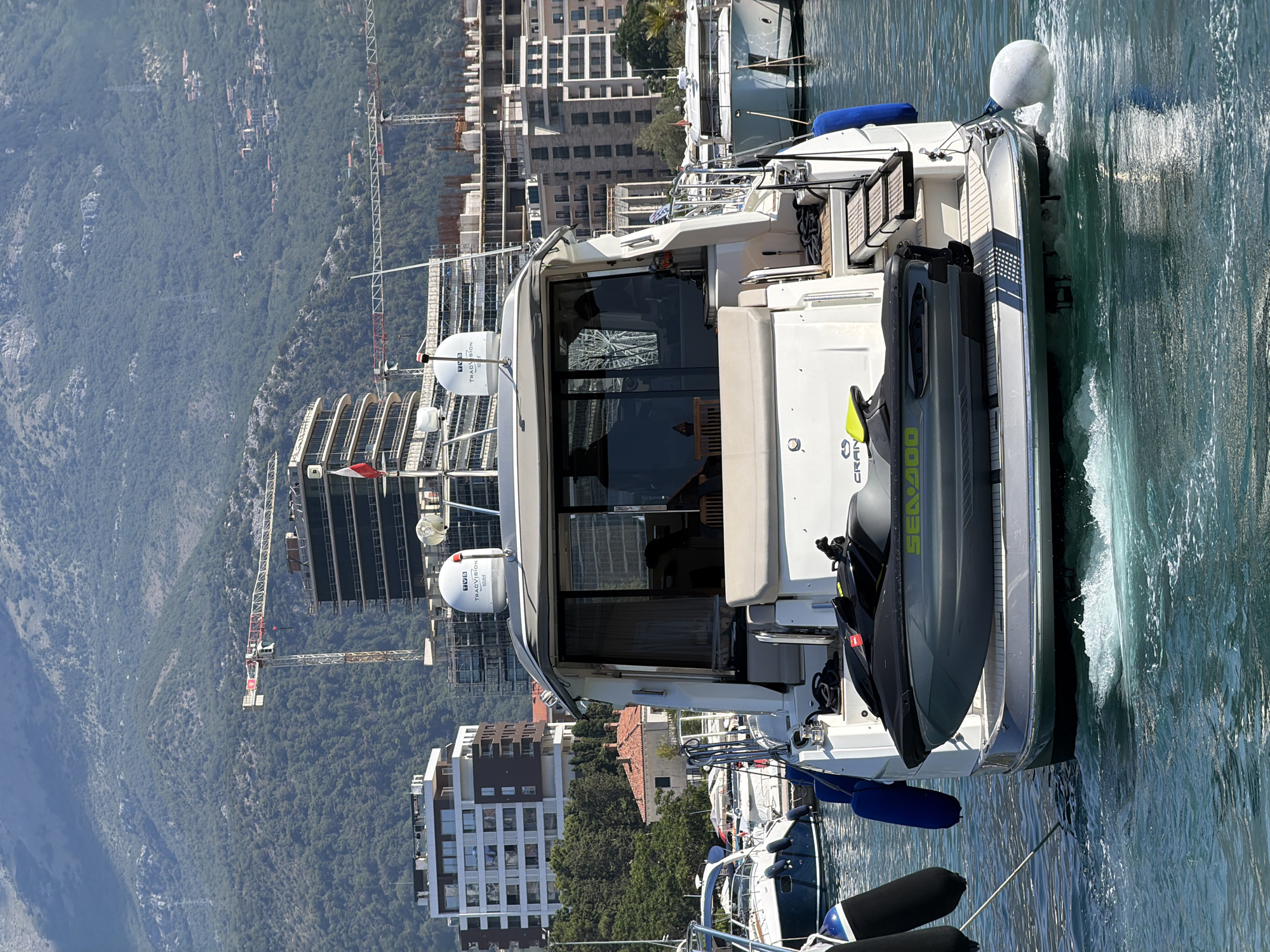 This image depicts the rear of a white motor yacht with a jet ski secured to its swim platform. The yacht is at anchor in a harbor, with other vessels visible in the background. The harbor is situated in front of a backdrop of lush green mountains, with modern buildings and construction cranes dotting the shoreline. The water is clear and blue, with some choppy waves visible around the yacht's stern. The scene appears to be a sunny day, with bright daylight casting reflections on the water and the vessels. The presence of construction suggests ongoing development in the area. The location is the Bay of Kotor, near the city of Budva, Montenegro, which is known for its picturesque coastline and developing infrastructure.