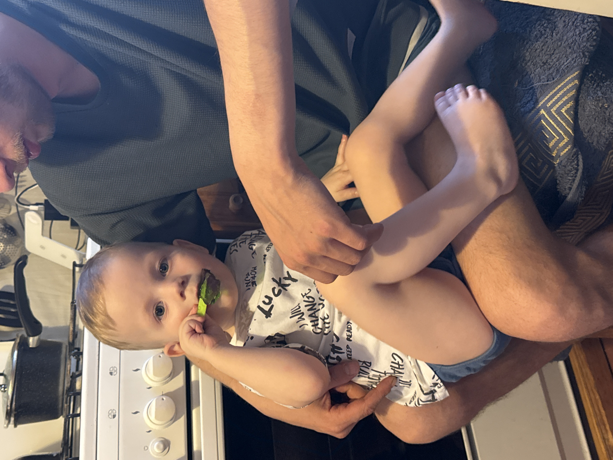 The image, taken in Saint Petersburg, Russia, shows a man holding a young child on his lap in what appears to be a home setting, possibly a kitchen or dining area. The image is oriented sideways, with the subjects appearing upright if rotated 90 degrees counter-clockwise.
The man, with light facial hair, is wearing a dark blue or grey waffle-knit top. His strong arm is visible, supporting the child. He is looking down and to the left, partially out of frame.
The young child, with fair skin, light hair, and blue eyes, sits on the man's lap. The child is wearing a white t-shirt with black text and light blue shorts or underwear. A green, mushy substance, likely food, is smeared around the child's mouth and on a bright green spoon the child is holding up towards their mouth. The child is looking directly at the camera with a neutral, slightly curious expression.
In the background to the left, a white stove or cooktop with black control knobs is visible, with a black pot and lid on one of the burners. Underneath the child's feet and legs, there is a dark blue or grey textured blanket or towel with a gold geometric pattern. Some electronic devices and cables are faintly visible near the man's side.
Visible text on the child's t-shirt includes words like "LUCKY," "WILL CHANGE," "KEEP READY," and "MORE FUN." The scene captures a quiet, everyday moment of a parent feeding a child.