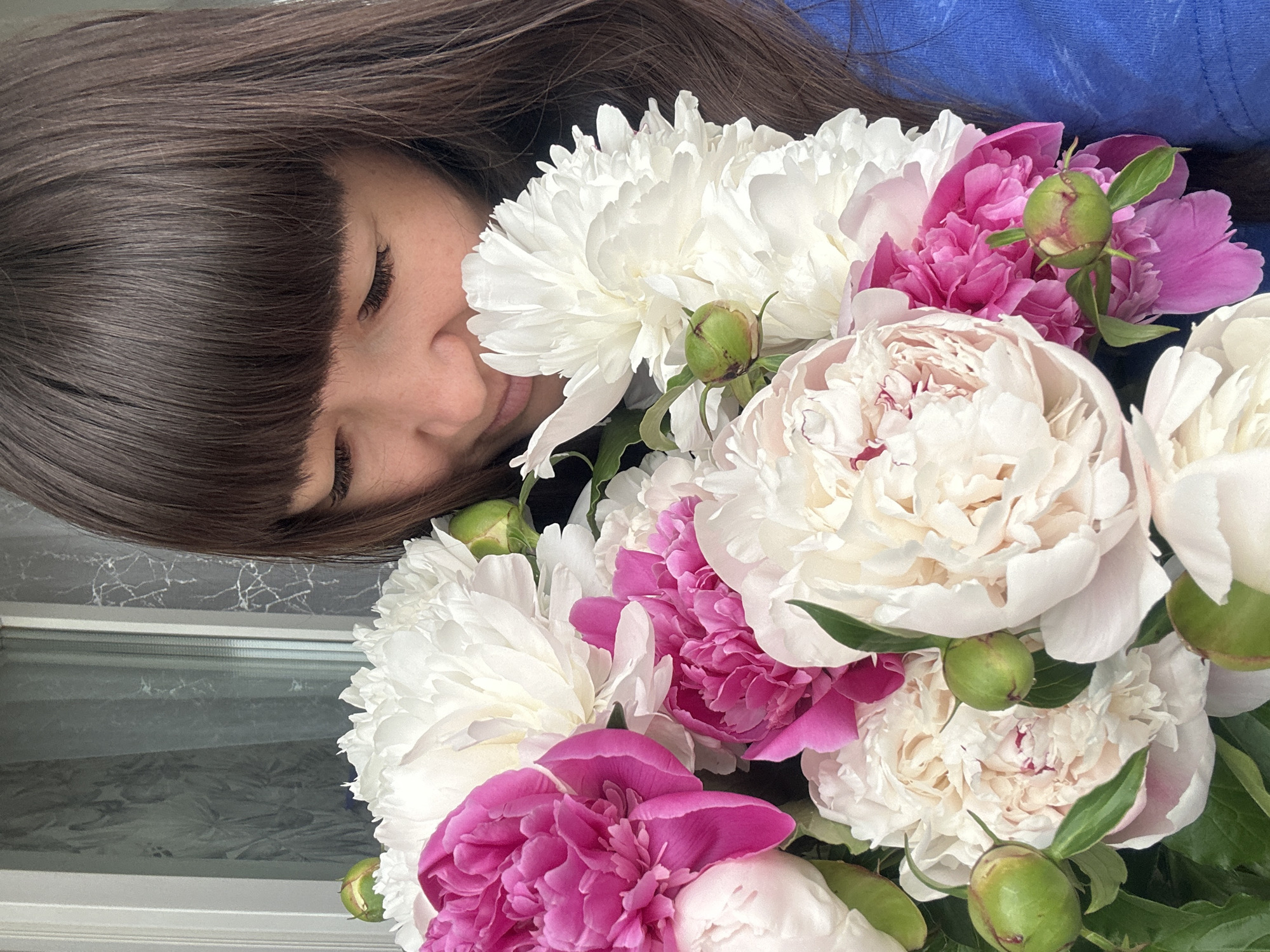 A woman with brown hair and bangs is holding a bouquet of white and pink peonies. She is wearing a blue top. The background is blurry, but appears to be a window. It is not possible to ascertain the location in the city of Chaikovsky, Russia from the image.