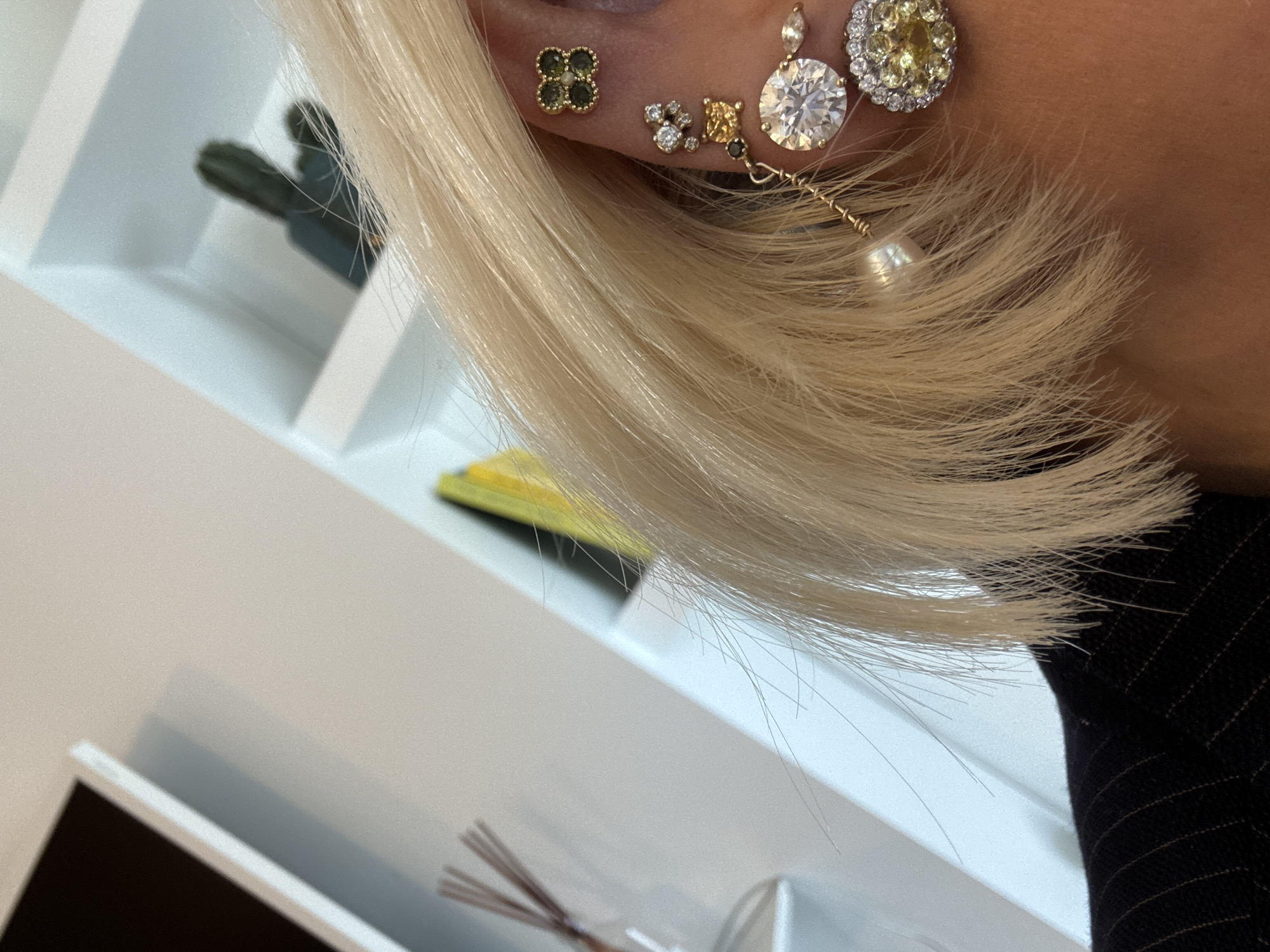 The scene captures a close-up, angled view of the right ear and adjacent scalp of an individual, positioned indoors. The individual has short, platinum-blonde hair partially obscuring the ear. The ear is extensively pierced and adorned with multiple earrings. From the upper cartilage, a gold-toned four-leaf clover stud with four green gemstones is present. Below this, a cluster of three small, clear, brilliant-cut stones is visible. Further towards the lobe, a square-cut yellow gemstone stud is worn. The earlobe prominently features a large, round, brilliant-cut clear gemstone set in a gold-toned bezel. Extending from this earring, a delicate gold-toned chain descends, terminating in a single white spherical pearl. Adjacent to the large clear stone, a circular earring with a central yellow gemstone surrounded by smaller clear gemstones is visible. A small, black, brilliant-cut stone and a marquise-cut clear stone are also present in this cluster. The individual's shoulder is visible, clad in a dark garment with light-colored pinstripes. The background consists of a bright white wall and shelving units. Visible objects in the background include a dark rectangular screen or framed item, a glass diffuser bottle with dark reeds, a dark, segmented decorative item on a shelf, and a bright yellow rectangular object. The location is identified as the City of London, United Kingdom.