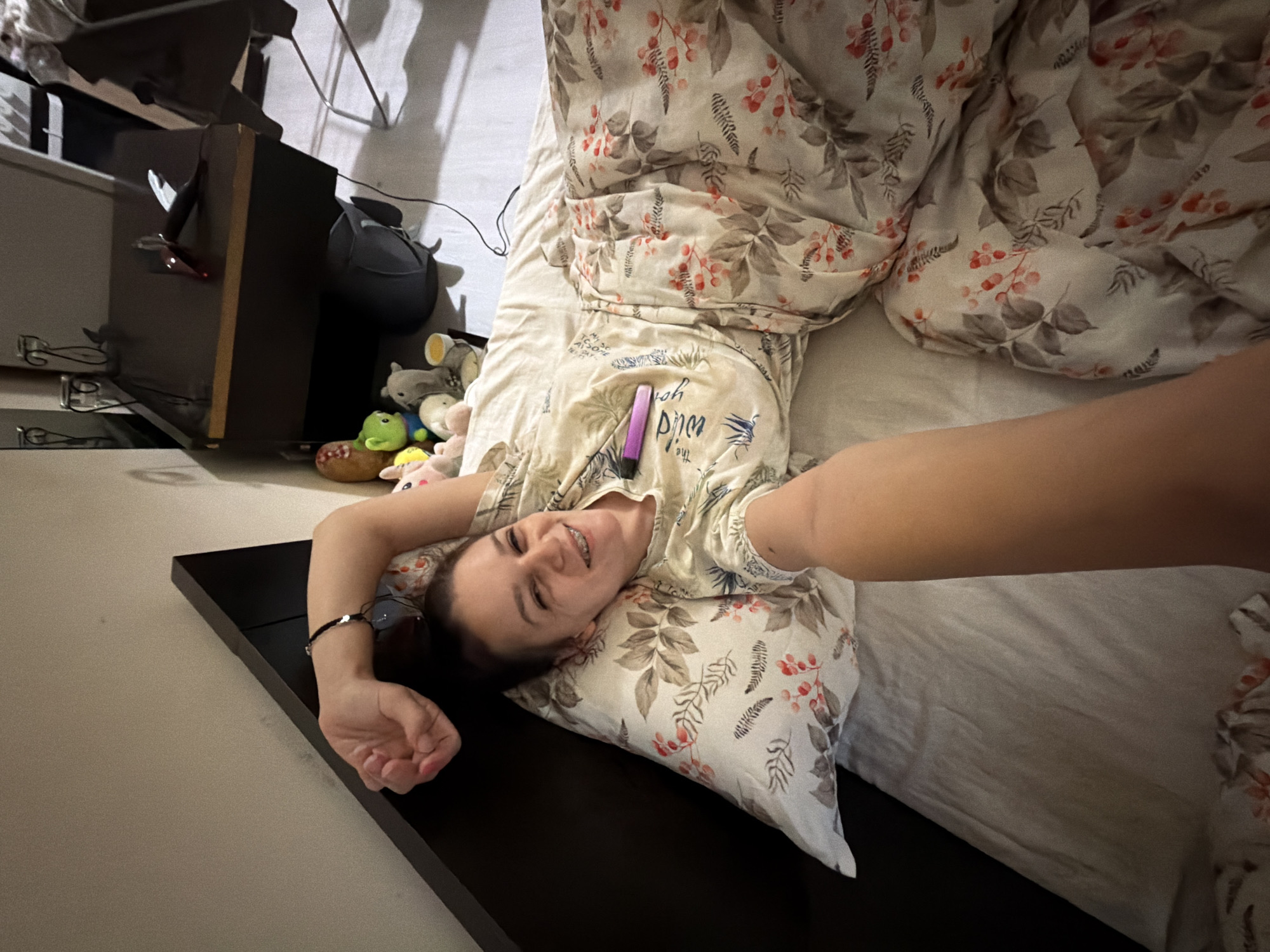 A woman lies in bed, holding a phone to take a selfie. She is wearing a light-colored shirt with a floral pattern and braces. Her right arm is extended upwards. The bed has a floral duvet and a white pillow. In the background, a nightstand holds various objects, including stuffed animals and a black speaker. The scene appears to be indoors, possibly a bedroom, in Sofia, Bulgaria.