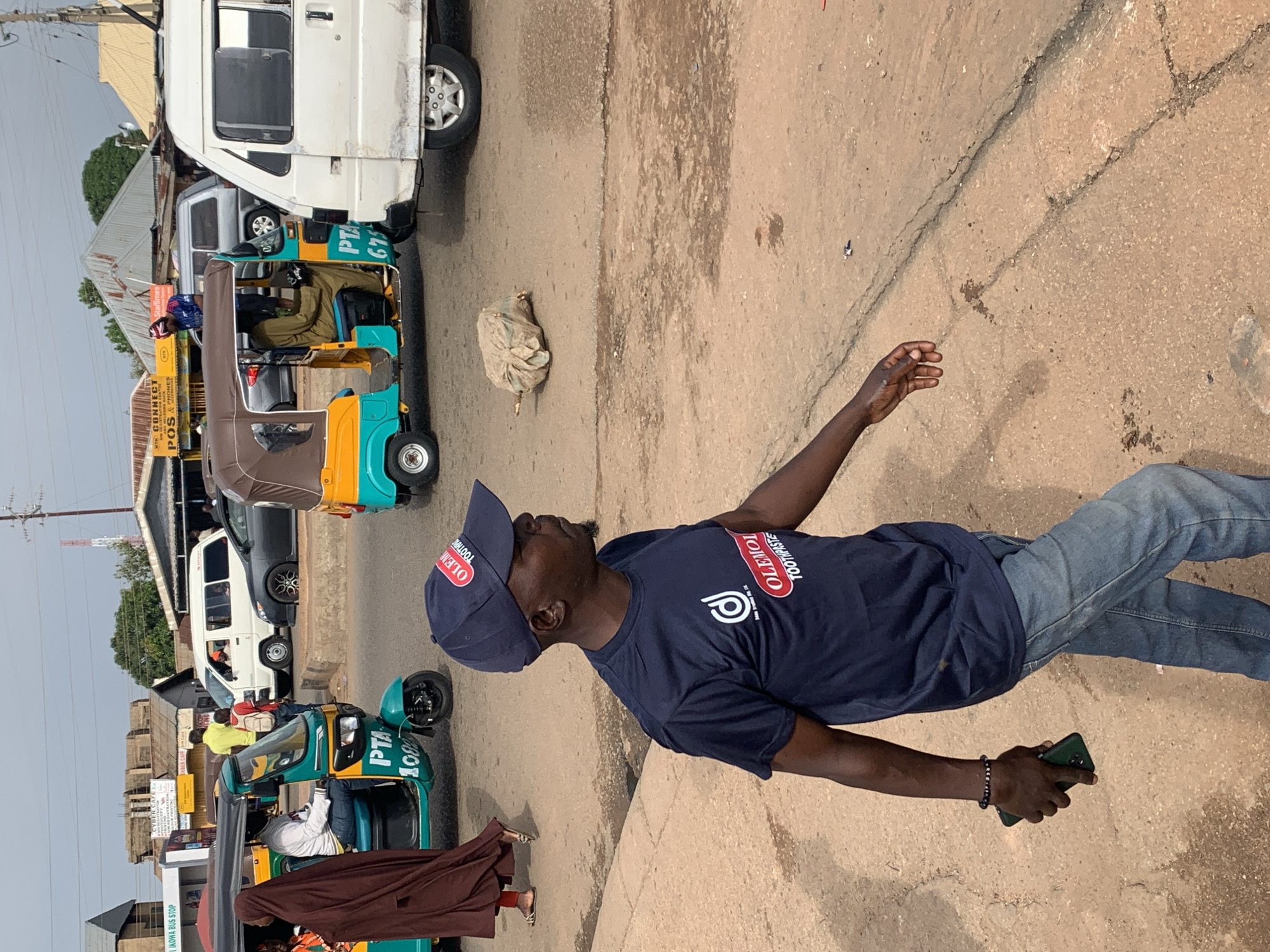 In Gura Top, Nigeria, an outdoor scene captures a busy street with several vehicles and pedestrians. In the foreground, a man wearing a navy blue t-shirt with white and red lettering and a matching baseball cap walks to the right, holding a dark smartphone in his left hand. His t-shirt has a circular logo on the left chest and the text "OLEMOOTHPASTE" across the front. He is also wearing light blue denim jeans.
Behind the man, a teal and yellow motorized tricycle, known locally as an "okada" or "tuk-tuk," is stopped. A person wearing a brown hijab and a dark brown garment is seated in the passenger area. The tricycle displays "PTA 1002" on its side. To the right of this tricycle, another similar tricycle, primarily yellow with a brown canopy, is also stationary. A person in a khaki-colored outfit is visible in the driver's seat.
Further back, a white van, possibly a public transport vehicle, is parked. A dark gray sedan is also present on the road. The background reveals buildings with corrugated metal roofs, signage for businesses such as "MTN CONNECT" and "CYBER CAFE," and utility poles with wires. The ground is a dirt and asphalt mix, showing signs of wear and cracks. The sky is overcast.