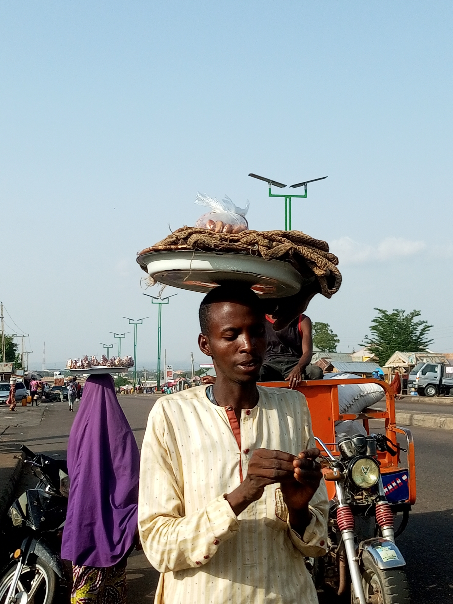 In Gombe, Nigeria, a man stands in the foreground, his head adorned with a large, shallow tray stacked with goods, including what appears to be fabric and a plastic bag of items. He is wearing a cream-colored, striped tunic. Behind him, a woman in a vibrant purple head covering and patterned skirt is also carrying a tray on her head. To the right of the man is an orange, three-wheeled motorcycle, commonly known as an "at Bartholomews," with a rider in a dark shirt seated on it. The scene unfolds on a paved road with vehicles and other pedestrians in the background, suggesting a busy urban environment. Streetlights with double solar panels are visible along the roadside, and in the distance, buildings and trees indicate a developed area. The sky is clear and bright, suggesting daytime.