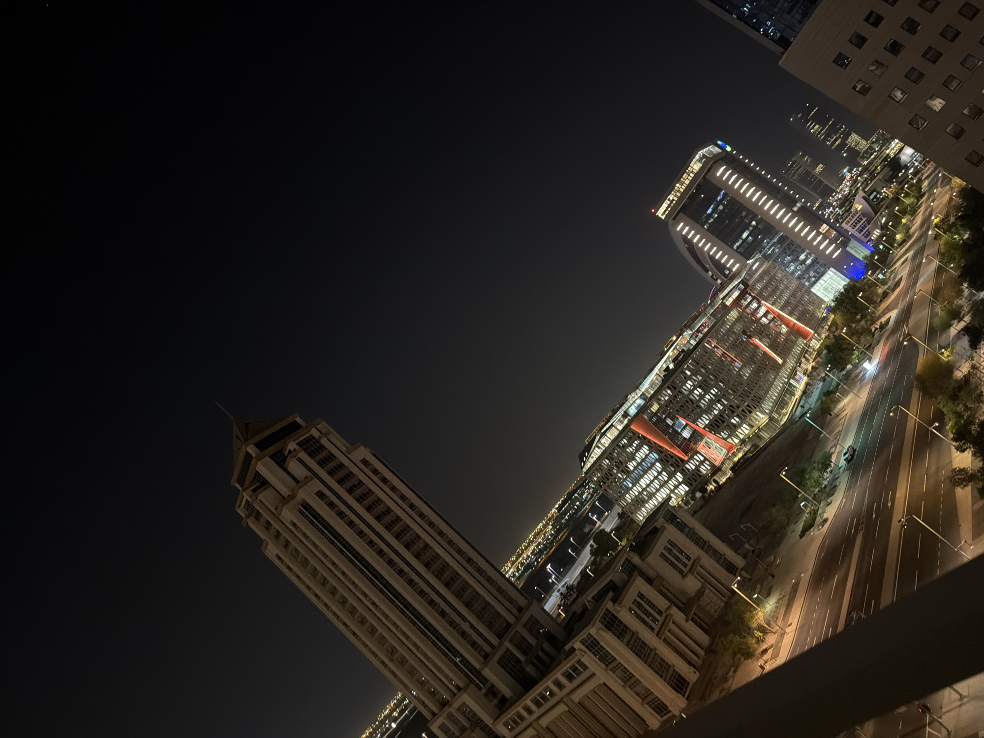 The image shows a nighttime aerial view of a modern cityscape. The perspective is from a high vantage point, looking down and to the right at a collection of illuminated buildings and a street below.
Dominating the left side of the frame is a tall, slender skyscraper with a distinctive pointed top, its facade featuring vertical lines and numerous windows. To its right and slightly lower, a cluster of buildings is visible, characterized by geometric designs and illuminated windows. One of these buildings has prominent red and orange vertical accents. Further to the right and in the mid-ground, another skyscraper stands out with a bright, white-lit facade. In the background, a dense cityscape stretches into the distance, with numerous lights indicating more buildings and activity.
Below the buildings, a multi-lane road is clearly visible, with streetlights lining its edges. A few cars are present on the road, and streetlights illuminate the asphalt. Trees and greenery are sparsely distributed along the sides of the road. The overall impression is one of a vibrant, well-developed urban environment at night. The sky is dark and clear, with no visible celestial bodies.