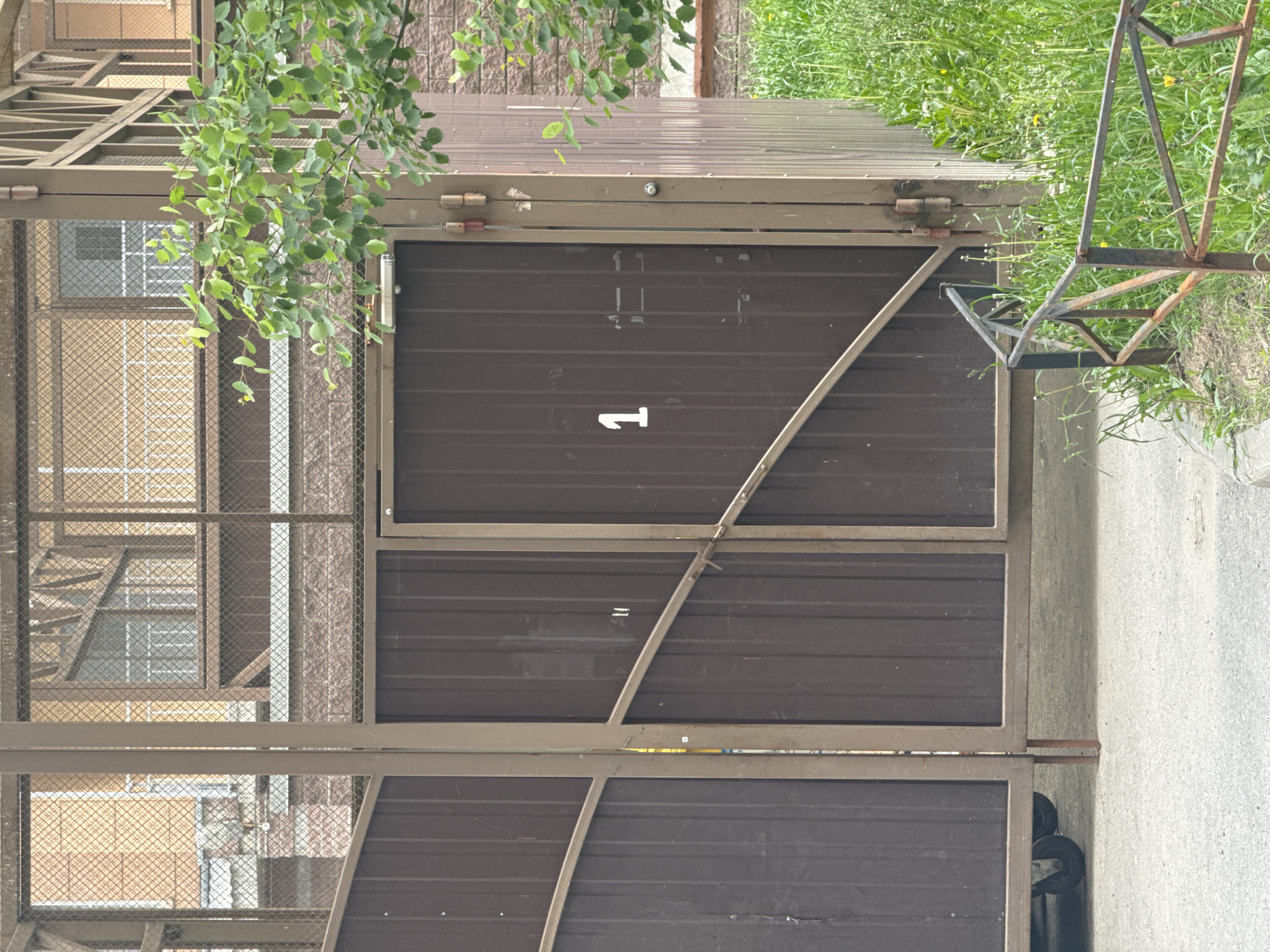 The image displays an outdoor scene featuring a robust metal structure, likely a storage unit, utility shed, or part of a car wash, under what appears to be an overcast daytime sky. The primary subject is a large gate or door made of brown corrugated metal panels, set within a lighter-colored metal framework.
A prominent white number "1" is stenciled on the upper right section of the gate. A diagonal metal brace crosses the gate from the upper left to the lower right. The gate is secured by hinges visible at its top.
The larger structure incorporates additional panels, some solid corrugated metal and others featuring a wire mesh design. Through these mesh sections, the background reveals parts of a building with brick walls and windows, characterized by white frames and gridded panes.
Overhanging the structure from the left are branches laden with vibrant green leaves, suggesting the presence of a tree or large shrub. To the right of the metal structure, a patch of bright green grass is visible, alongside a rusty, angular metal frame, possibly a stand or part of an old fence. The ground in front of the structure is composed of grey concrete. Partially obscured dark, circular objects, possibly wheels or equipment, can be seen behind the lower section of the gate.
The scene is static, devoid of people or direct activity, presenting a functional, utilitarian area.