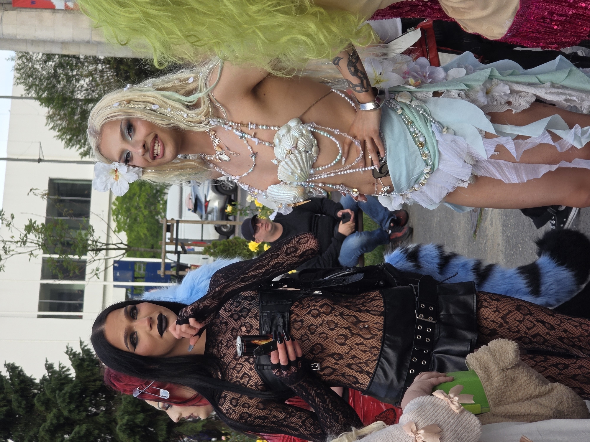 The image shows a group of people outdoors, possibly at a parade or festival, in Warsaw, Poland. In the foreground, two individuals are prominently featured. The person on the left has long black hair, dark eye makeup, and black lipstick. They are wearing a black lace top with a black leather bustier, black fishnet stockings, and a black leather skirt. They are holding a silver can and a black object. A blue and black furry tail is visible behind them.
The person on the right has blonde hair with a white flower tucked behind their ear and accessorized with beads and seashells. They are wearing a seashell bra and a light blue and white layered skirt adorned with shells and flowers. They are also wearing multiple necklaces and a silver bracelet.
In the background, a man wearing a black baseball cap and glasses is sitting on a bench. Other people and buildings are visible, along with trees and some yellow flowers. The setting appears to be an urban environment, possibly a street or a park.