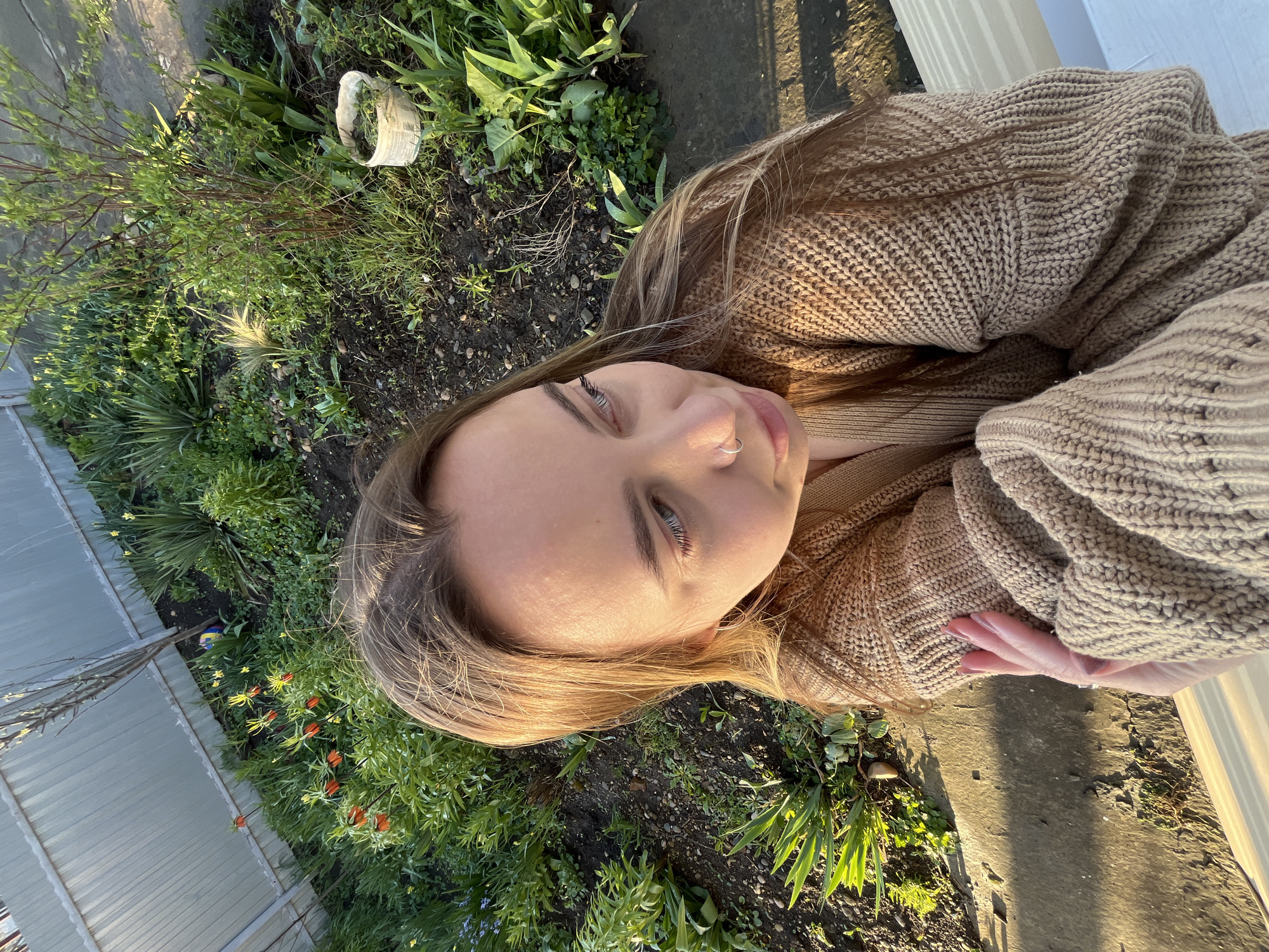 A young woman is lying on the ground next to a garden bed. She is wearing a beige knit sweater. The garden bed contains various plants, including some with orange flowers. The ground is covered in soil and greenery. In the background, a fence and other structures are visible. Based on the location provided, the image was likely taken in Novopokrovskaya, Russia.
