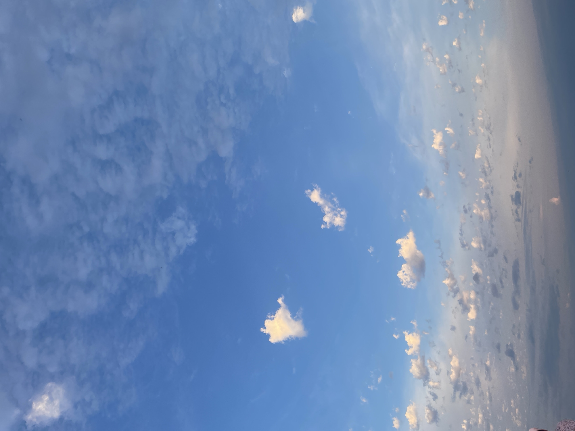 The image shows a wide view of a blue sky with scattered clouds. The clouds range in size and density, with some appearing as small, wispy formations and others as larger, more defined puffs. The lighting suggests it may be late afternoon or early morning, with some clouds tinged with pink and orange hues. The horizon is visible at the very bottom of the frame, appearing as a soft, hazy line of muted colors. There is a faint silhouette of a person on the left side of the frame, wearing a light-colored garment with a patterned design. The overall impression is one of a peaceful and expansive natural scene.