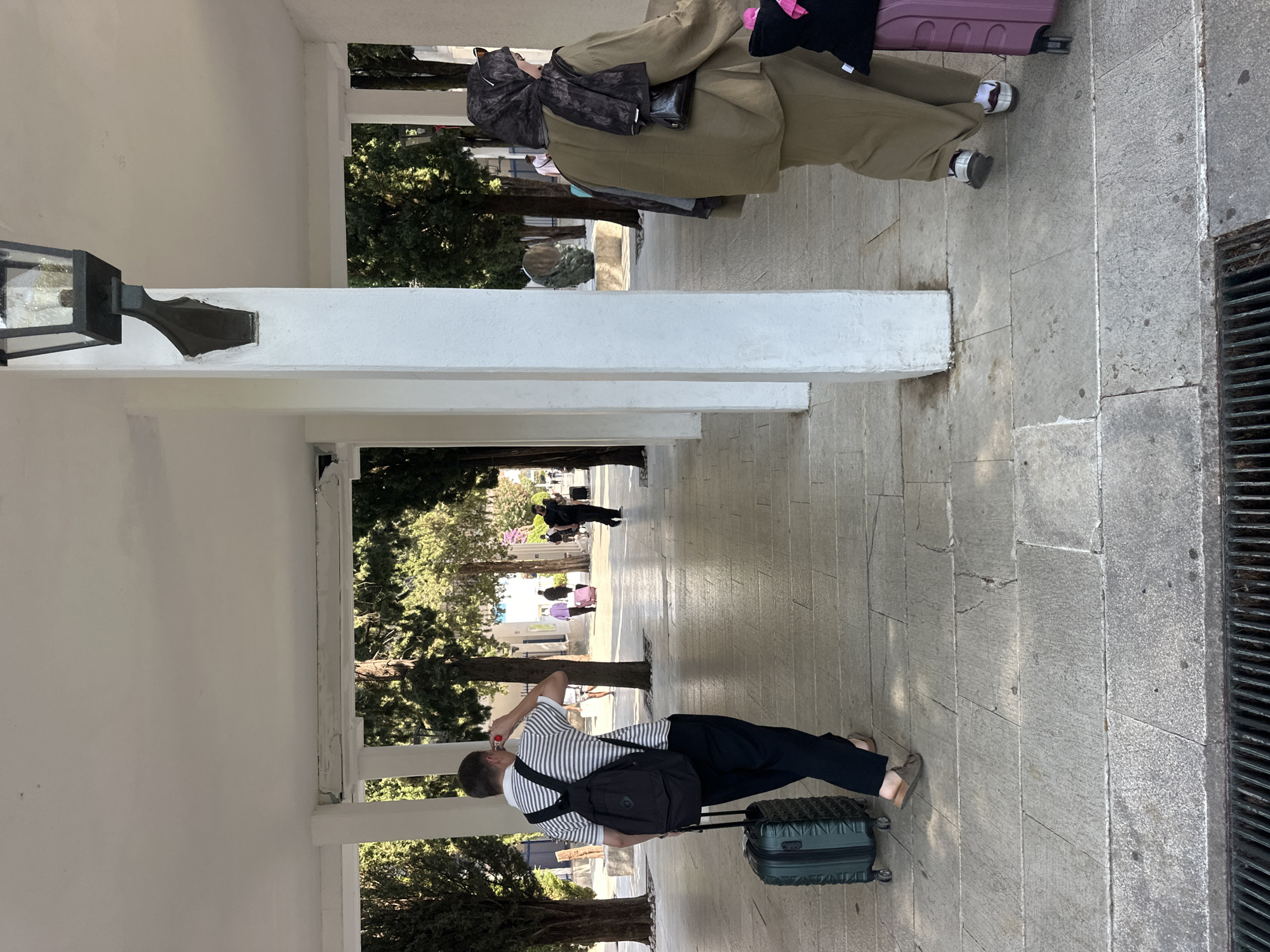 The image depicts an outdoor walkway, likely in Budva, Montenegro, with people engaged in travel. In the foreground, a man with a black backpack is pulling a green suitcase. Further down the walkway, a woman wearing a hijab is walking with a purple suitcase. The walkway is paved with stone slabs and is covered by a white overhang supported by pillars. Trees are visible in the background, suggesting an urban or park-like setting. The lighting indicates it is daytime.